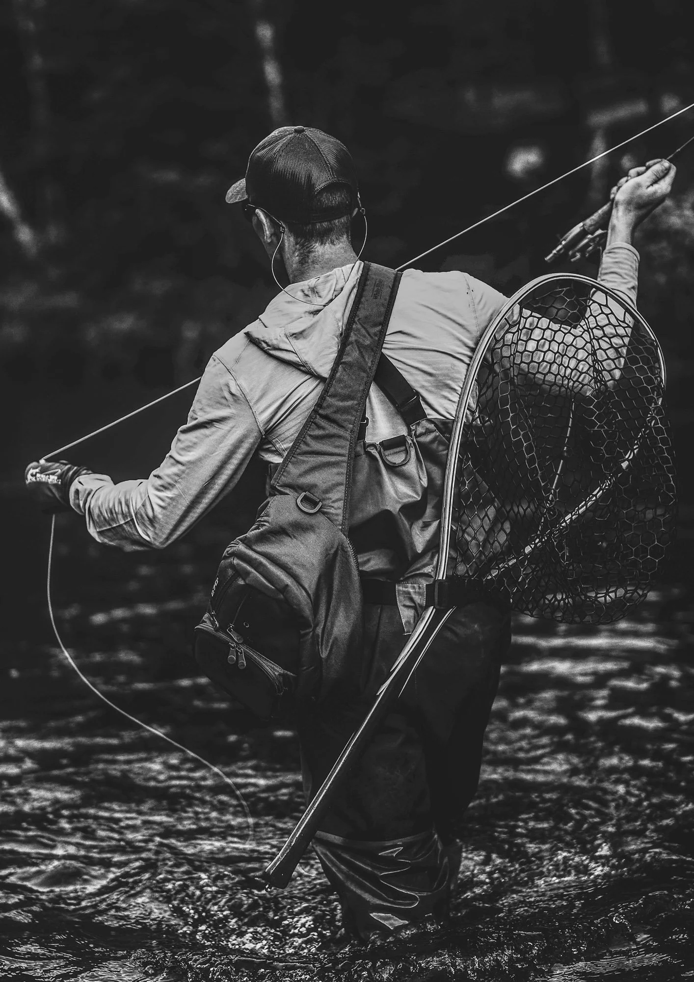 A person wearing a cap and outdoor gear stands in a stream, fishing with a rod and net, with water splashing around.