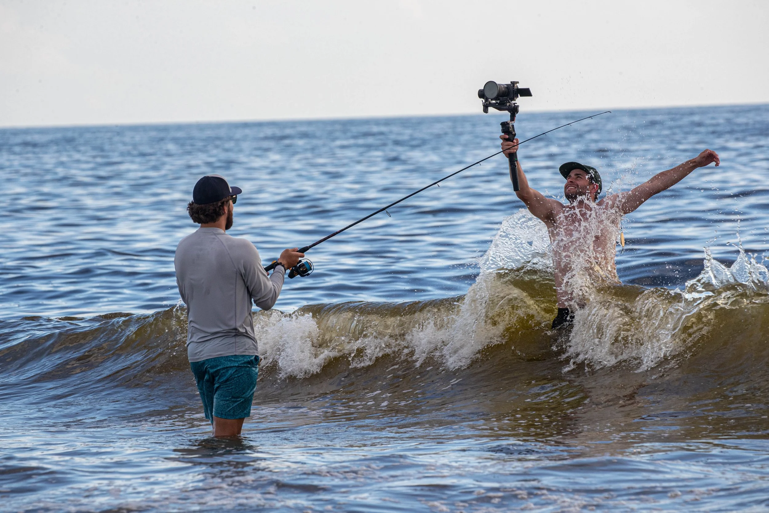 A man is in the water holding a camera on a stabilizer, taking a photo of another man who is jumping out of the water with a fishing rod in hand, at the beach.