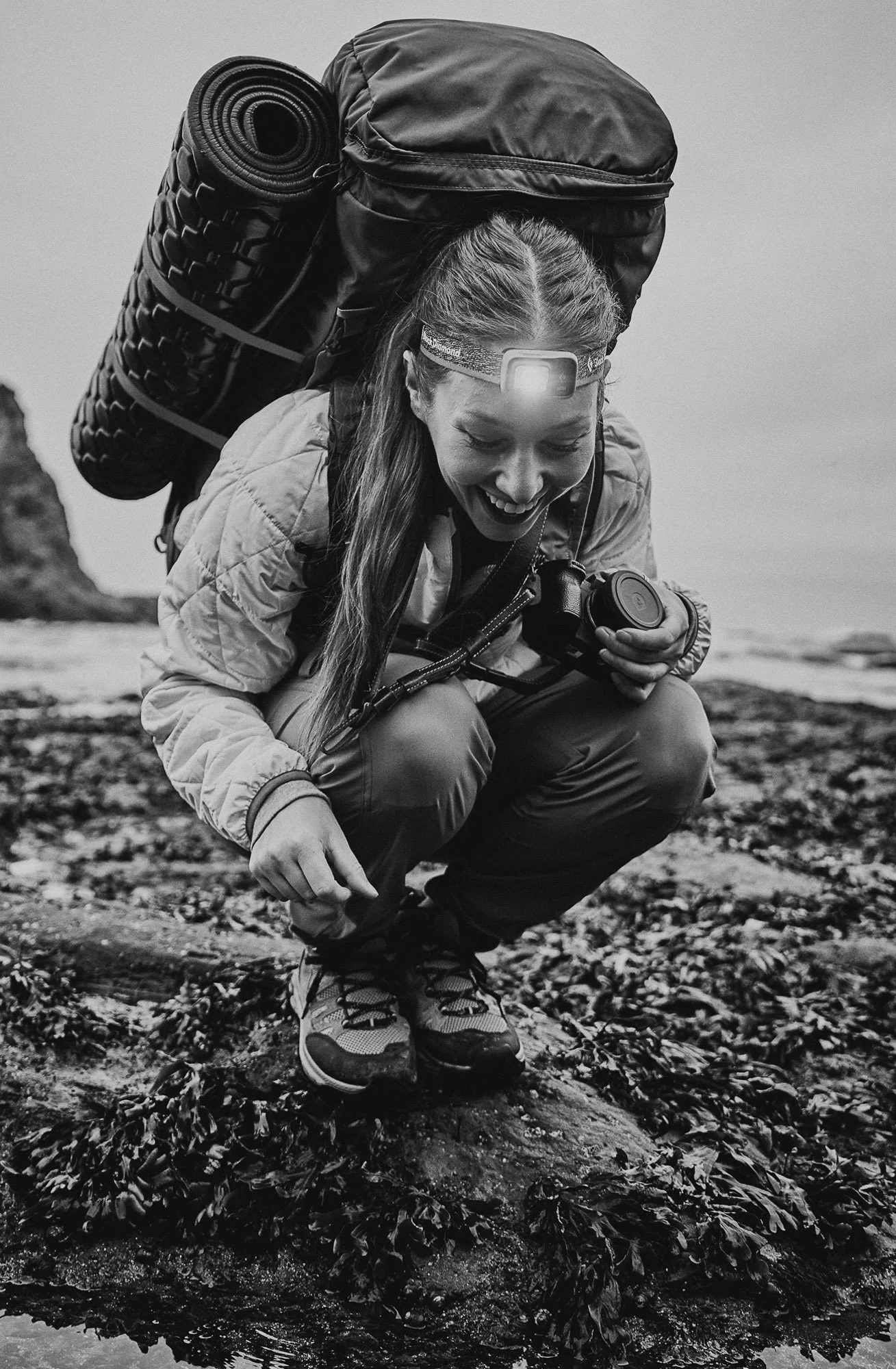 A woman crouching near a rocky shoreline with seaweed, holding a camera, wearing a headlamp and outdoor gear, with a backpack and sleeping pad on her back, smiling and looking down.