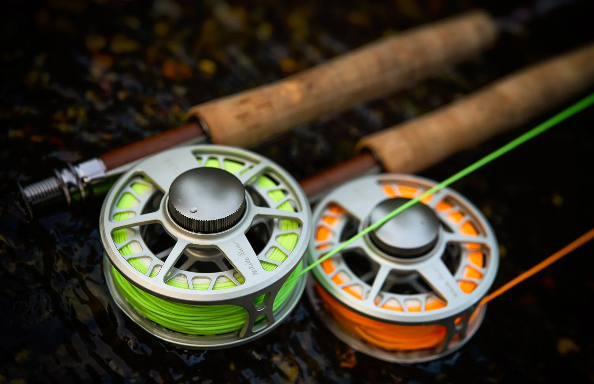 Close-up of three fishing reels on dark textured surface. The reels have green and orange fishing line wound around them, with two fishing rods in the background.