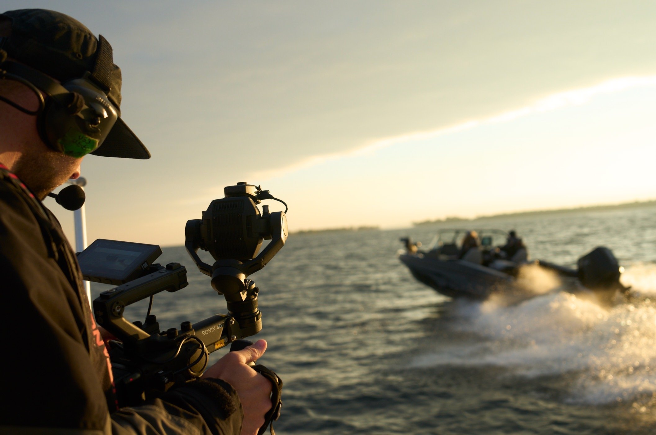 A person wearing a hat and headset operating a camera on a tripod on a boat, with other boats on the water during sunset.