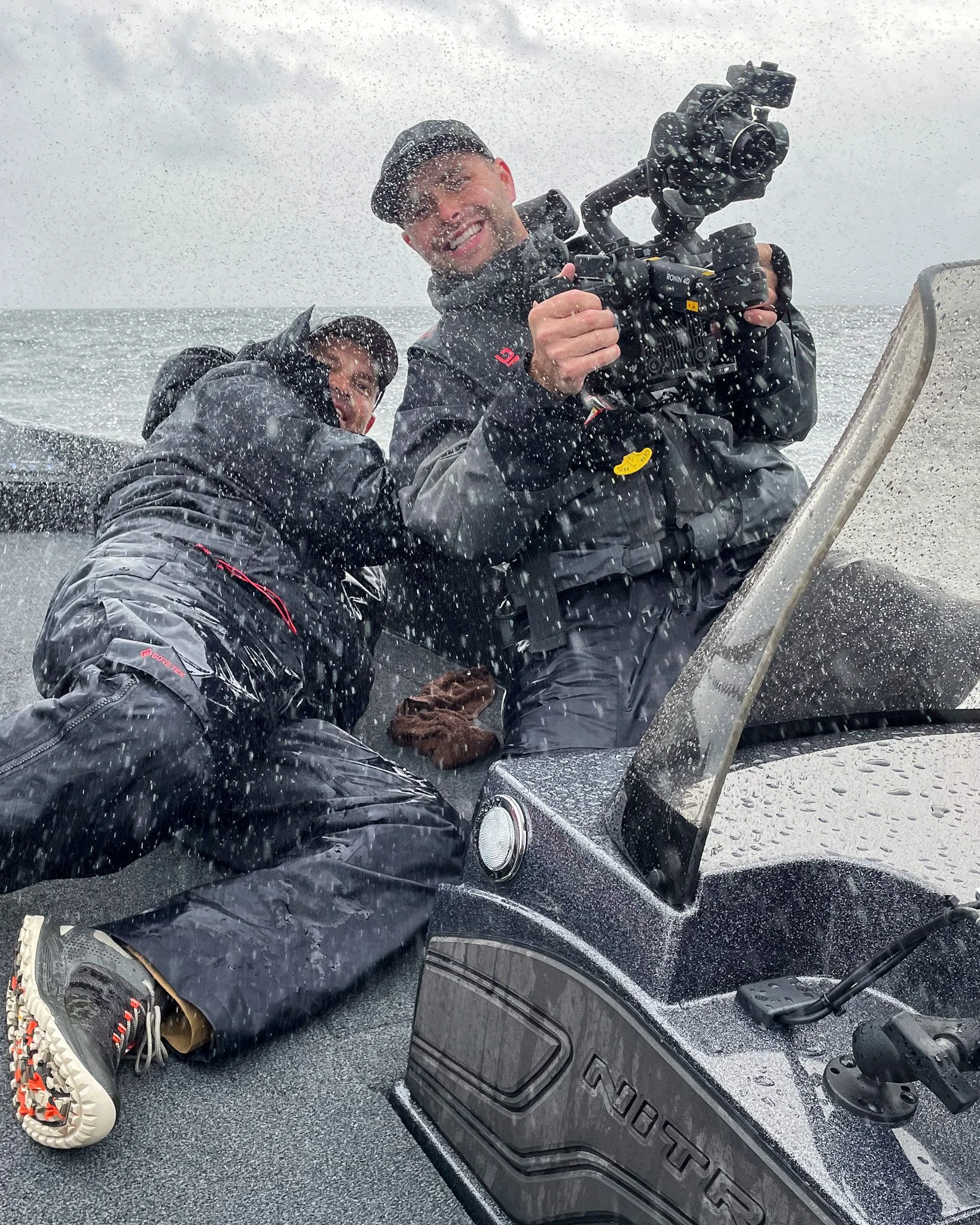 Two men are on a boat, with one holding a professional video camera. They are dressed in wet weather gear, and there are splashes of water around them. The man holding the camera is smiling, and the boat is near the water's surface, which appears to be rough with spray.