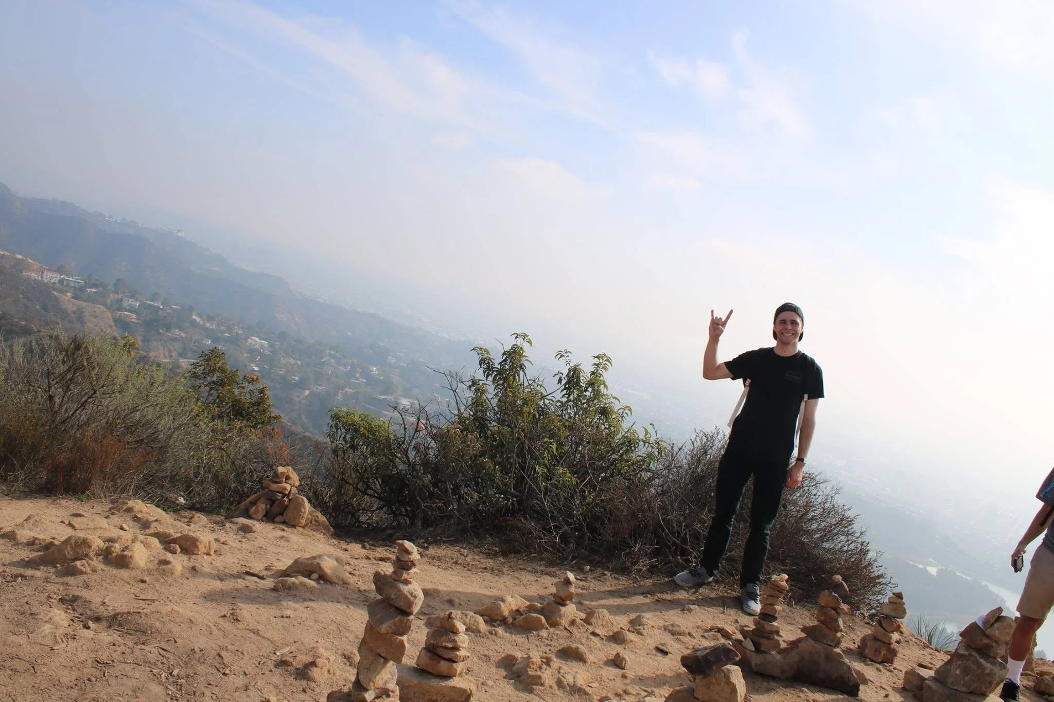 Person smiling and making a hand gesture in front of a hillside with stacked rocks and a view of the distant landscape.