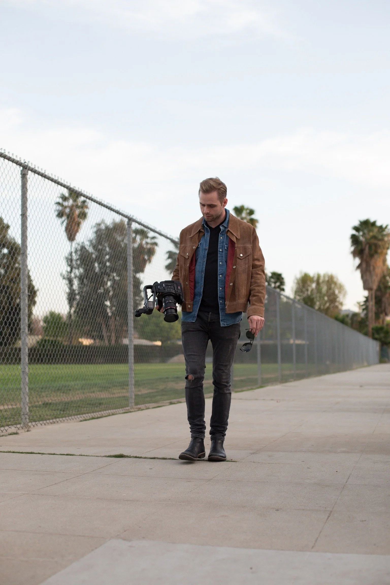 Young man walking on a sidewalk near a chain-link fence, holding a camera and sunglasses, with palm trees in the background under a partly cloudy sky.