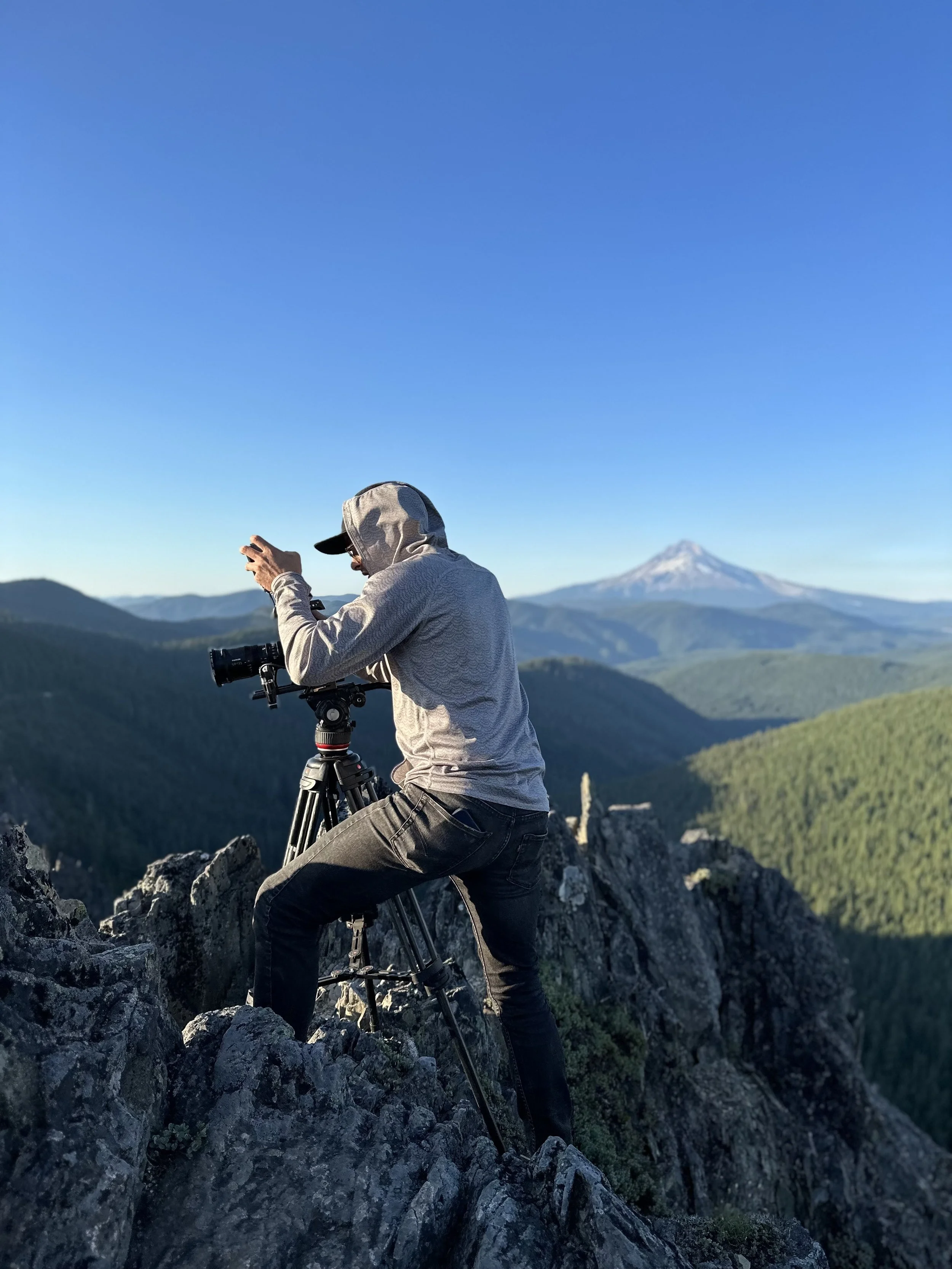 Person in gray hoodie and black pants taking a photograph with a camera on a tripod on rocky terrain, with forested mountains and a snow-capped mountain in the background under a clear blue sky.