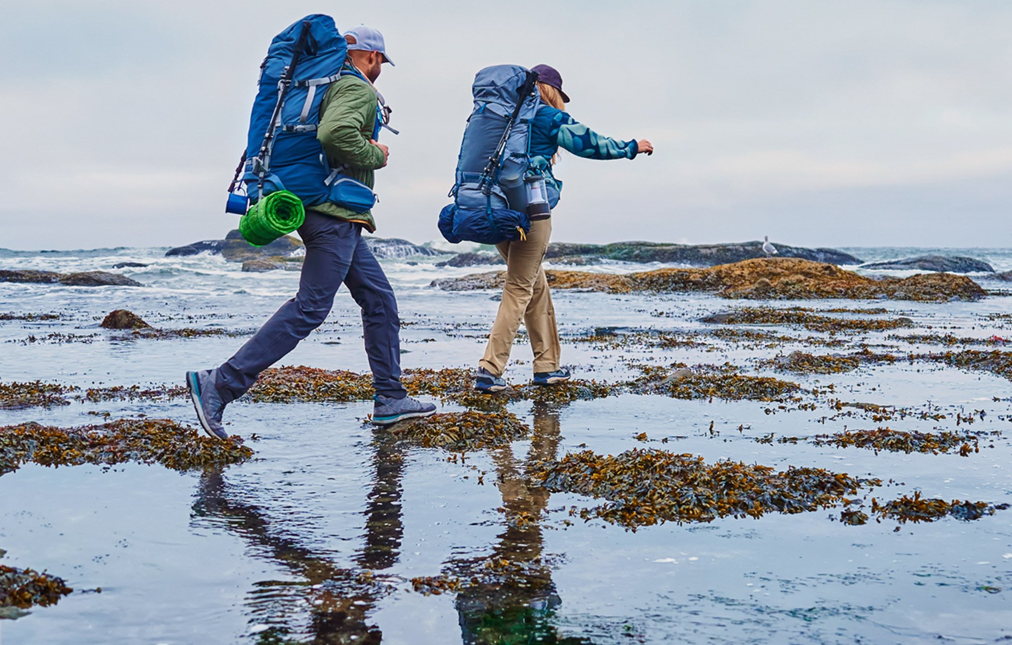 Two people hiking on a rocky beach with tide pools, both wearing backpacks and outdoor gear, with cloudy sky and ocean in the background.