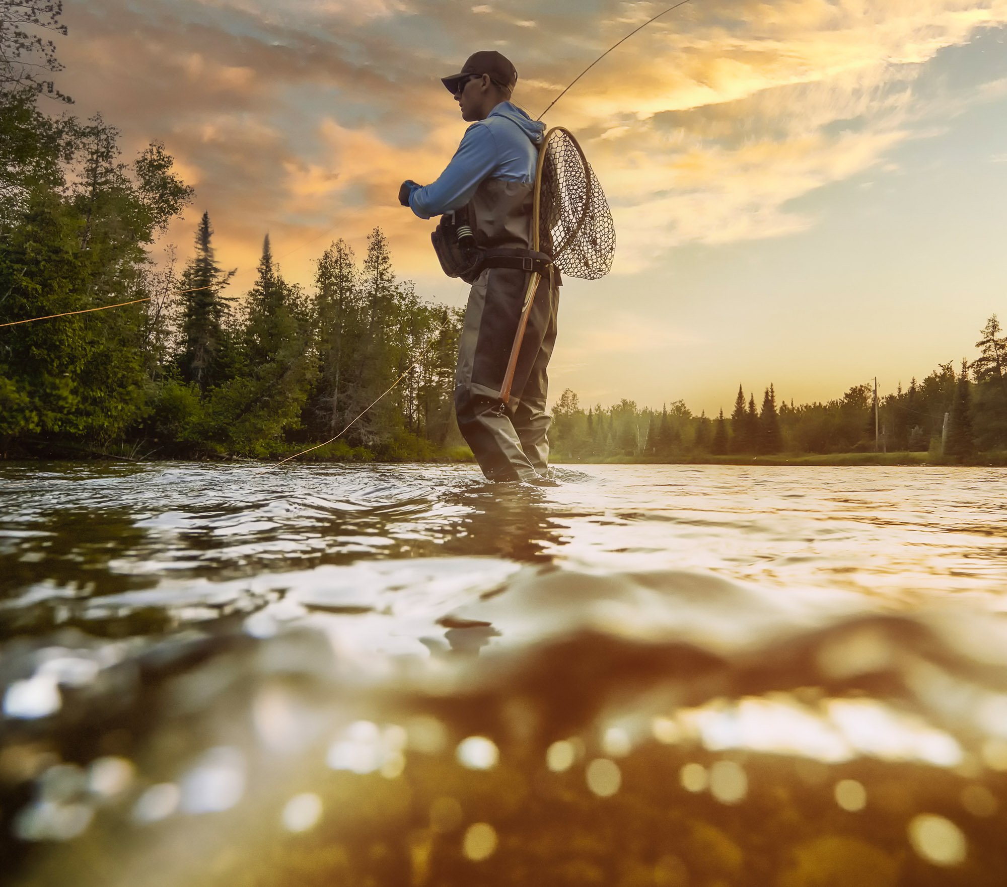 Person fishing in a river during sunset, wearing sunglasses and a hat, with trees in the background.