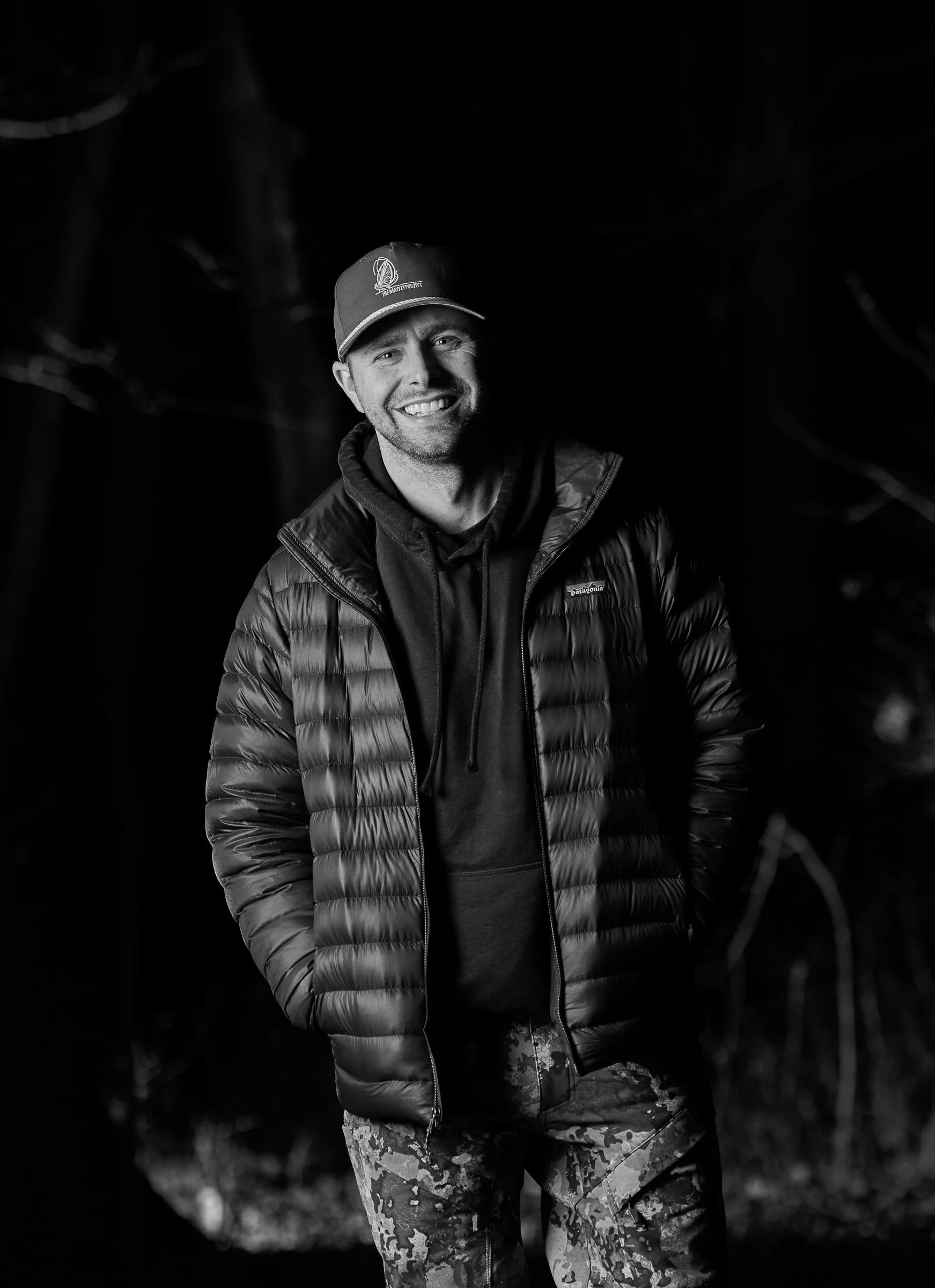 A smiling man wearing a baseball cap, puffy jacket, and camouflage pants, standing outdoors in a forest at night.