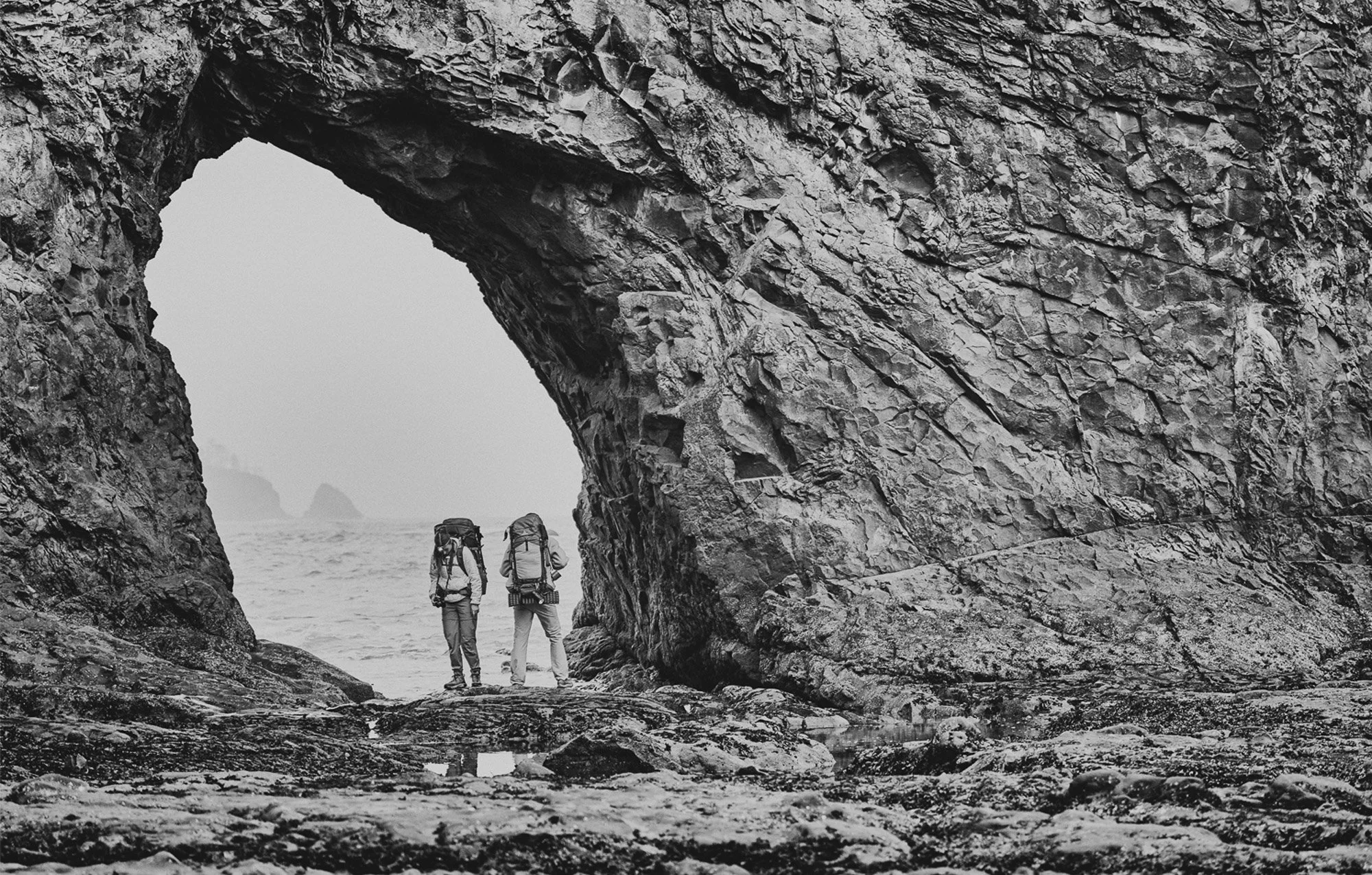 Three hikers with backpacks walk through a natural rock formation with a large archway, overlooking the ocean, in black and white.