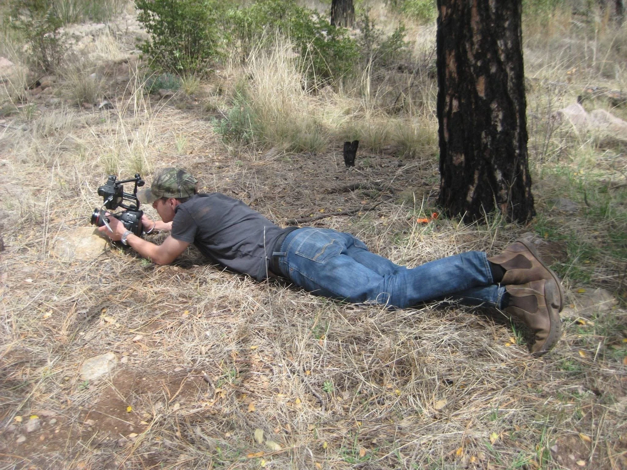 A person lying on the ground in a forest, holding a camera with a large lens and wearing a camouflage hat, with trees and dry grass surrounding them.