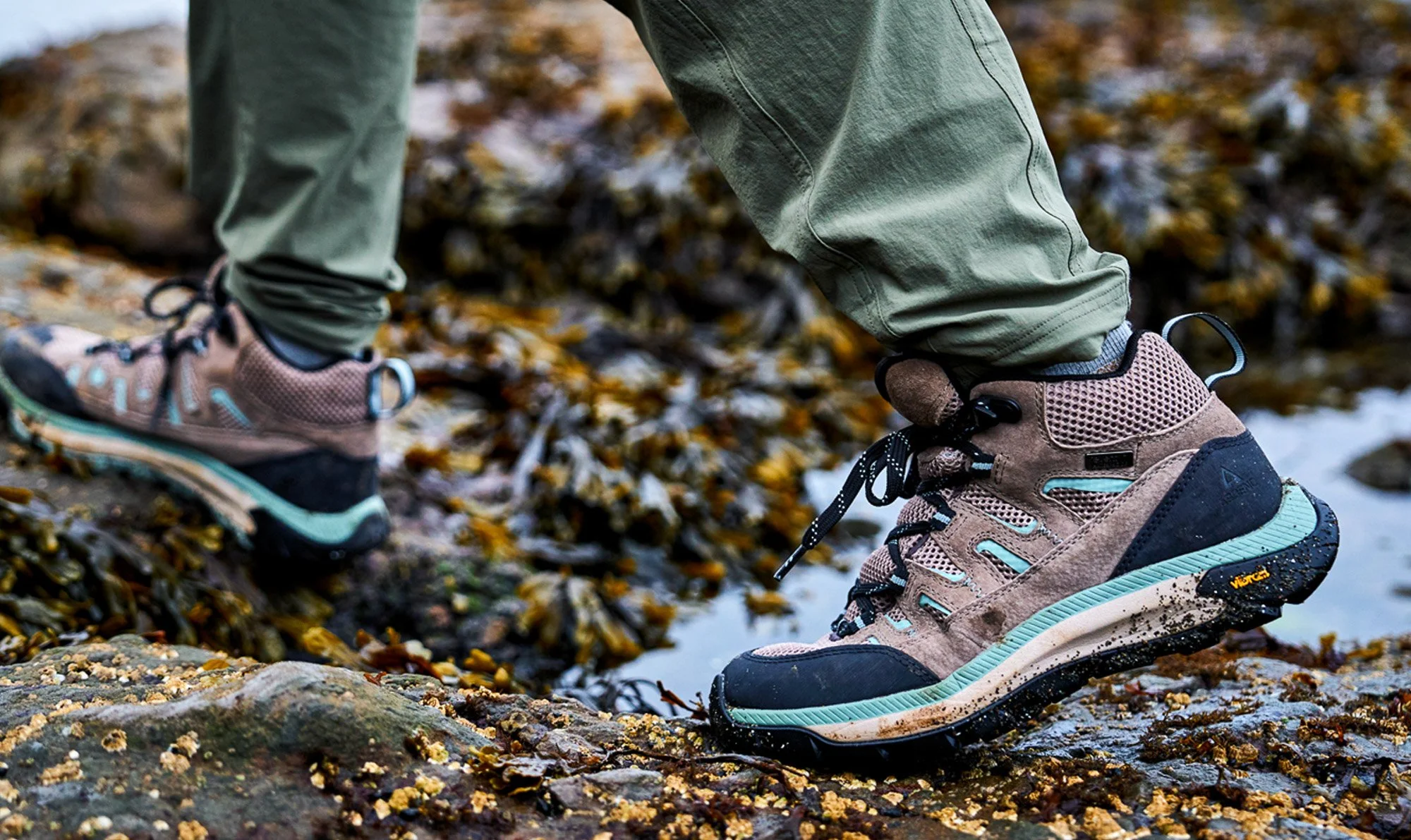 Close-up of a person wearing hiking boots and khaki pants walking on a rocky, leaf-covered surface near a small stream in an outdoor setting.