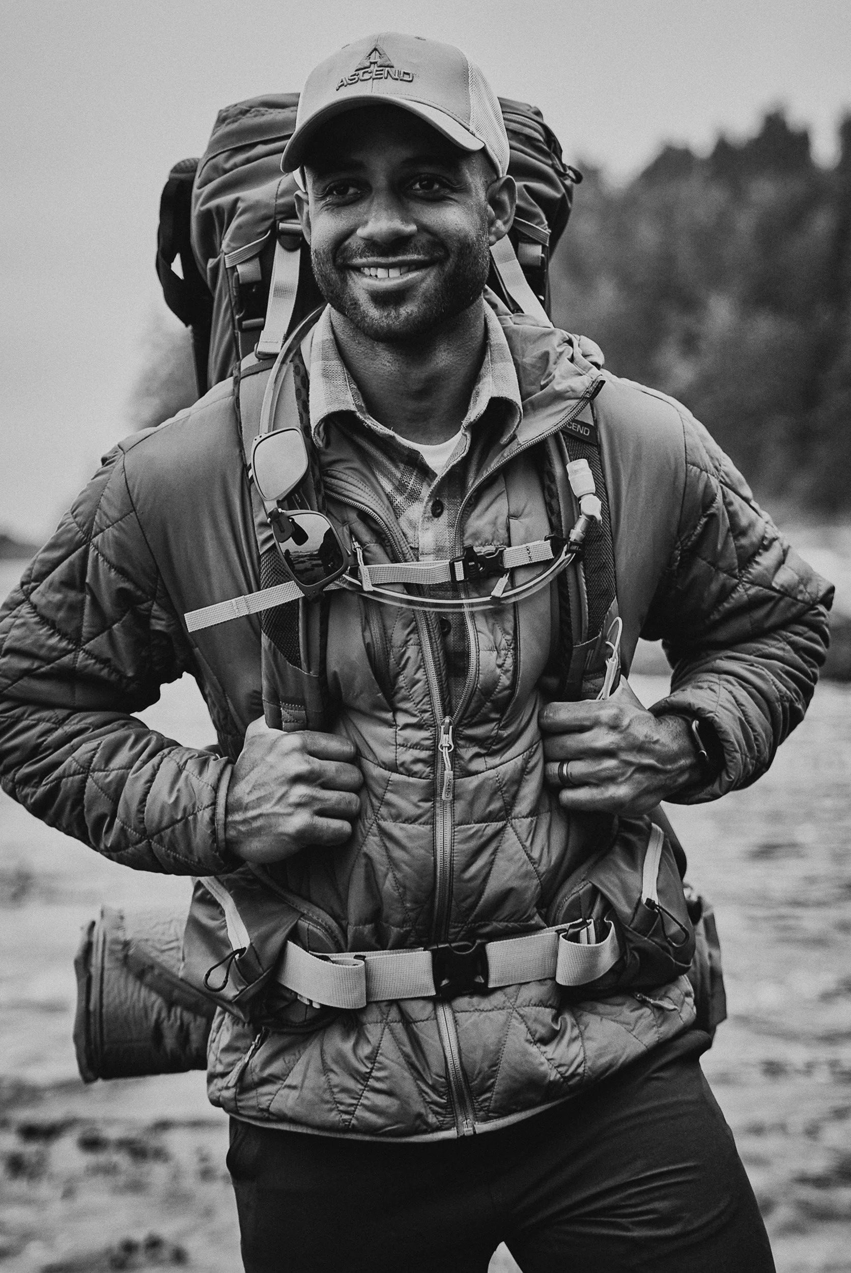 A man with a beard smiling, carrying a backpack, wearing a cap and outdoor gear, outdoors in a natural setting.