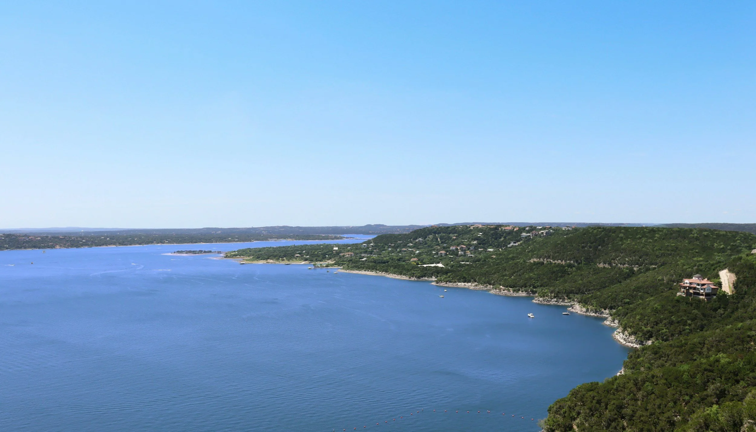 A large river with a few boats, surrounded by green hills and scattered houses, under a bright blue sky.