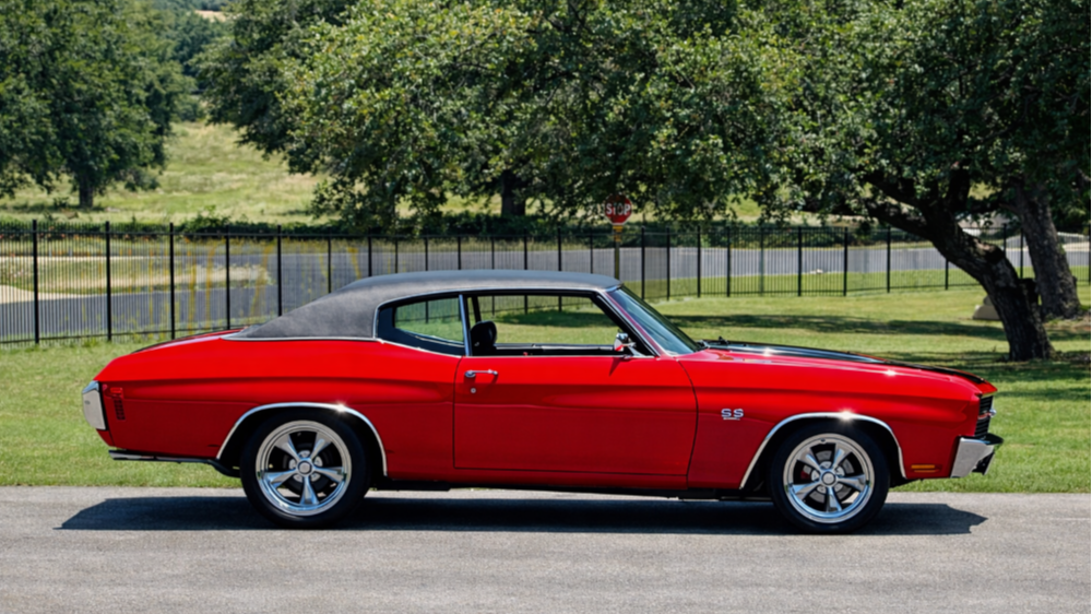 A red vintage Chevrolet Camaro SS convertible with a black soft top, parked on a street with trees and a fence in the background.