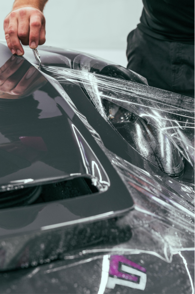 Person using a plastic pry tool to open a damaged black car hood with visible scratches and dents.