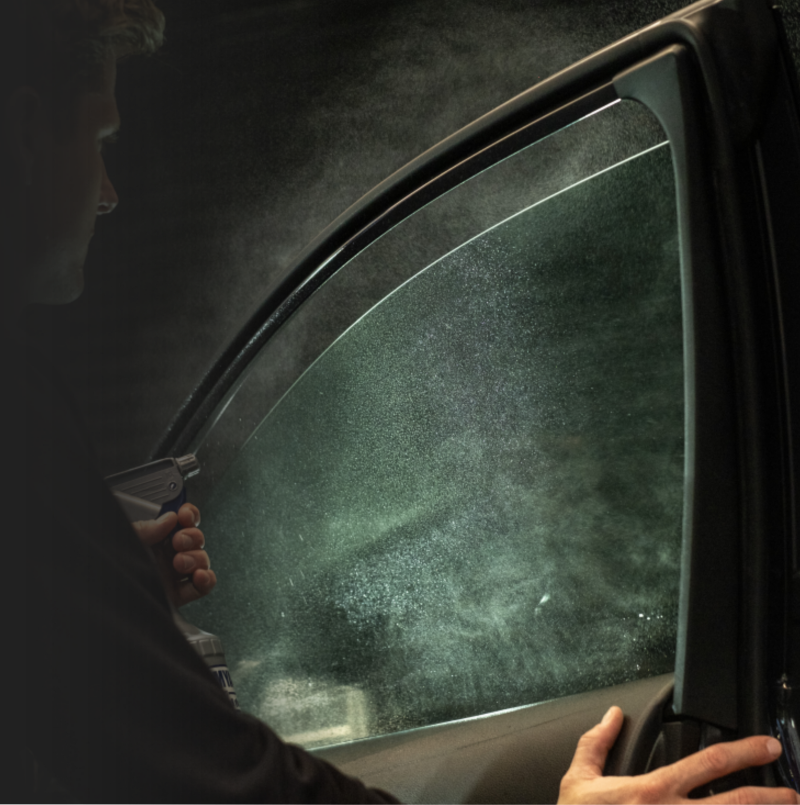 Person cleaning a car window at night, using a spray bottle and cloth, with rain visible on the window.