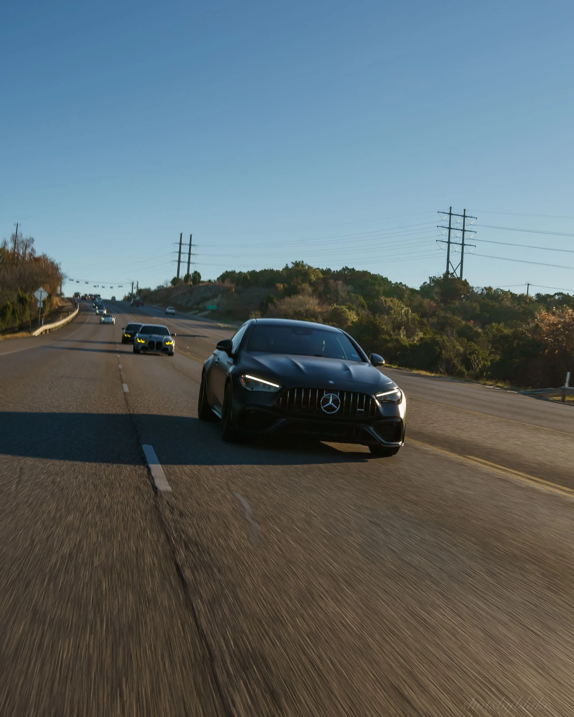 Black Mercedes-Benz car driving on a highway with several other cars in the background and a hill with trees and power lines under a blue sky.
