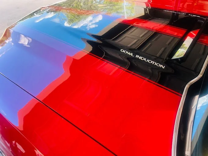 Close-up view of the front part of a colorful car, with a red, blue, and black hood, reflecting a partly cloudy sky and a building wall on its shiny surface.