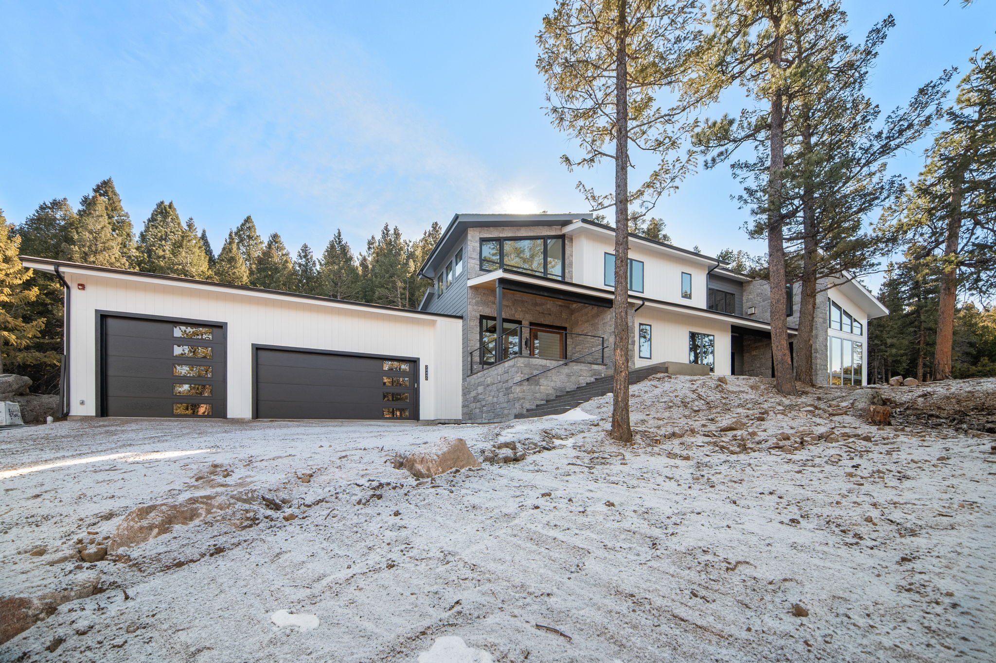 Modern multi-story house with large glass windows and a wraparound deck situated on a hillside with forested landscape.