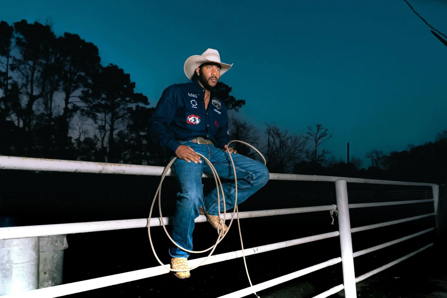 A man wearing a cowboy hat and western shirt sits on a white fence holding a rope, with trees and a dark sky in the background.