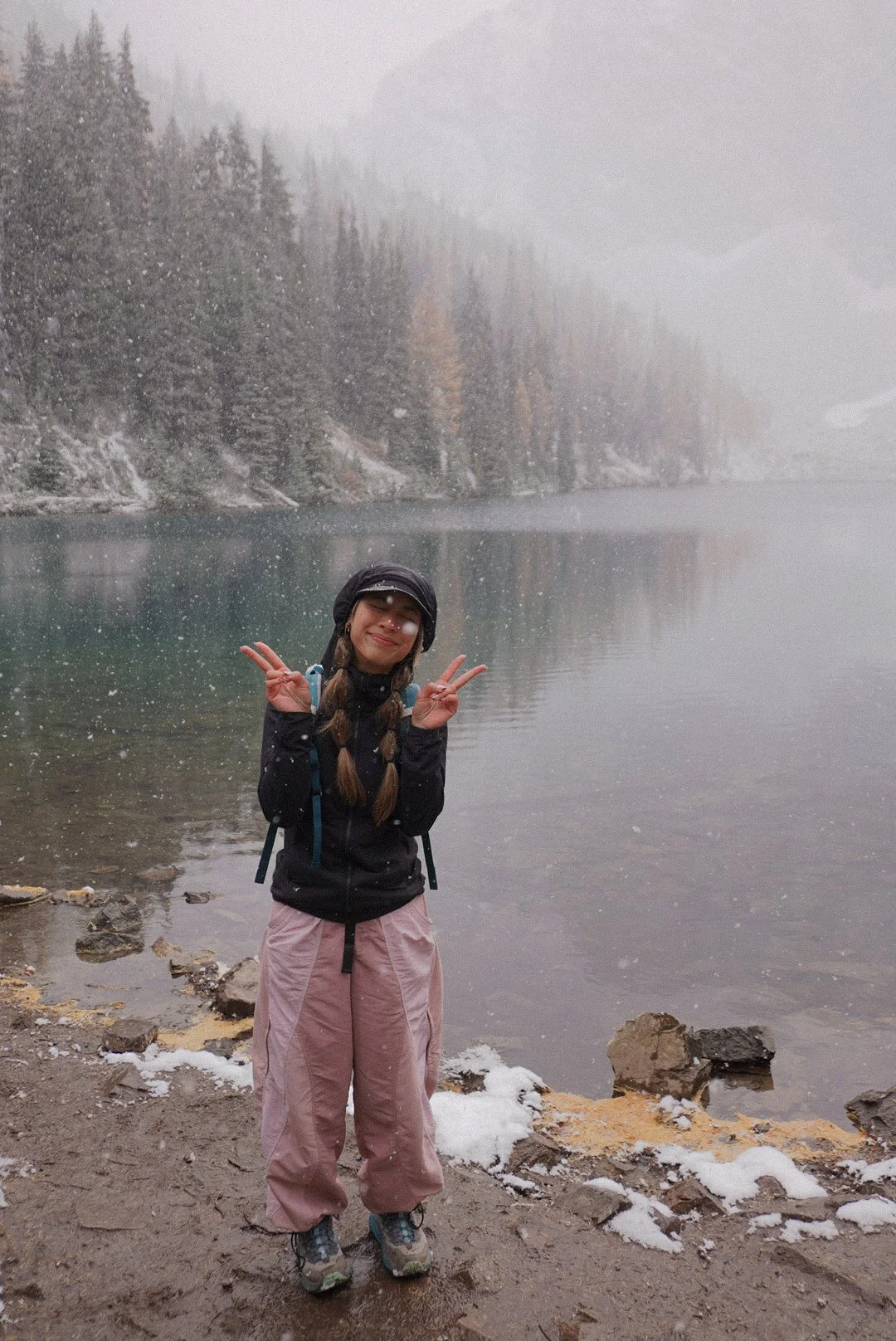 A woman stands by a lake in a snowy, foggy forest, smiling and making peace signs with both hands.