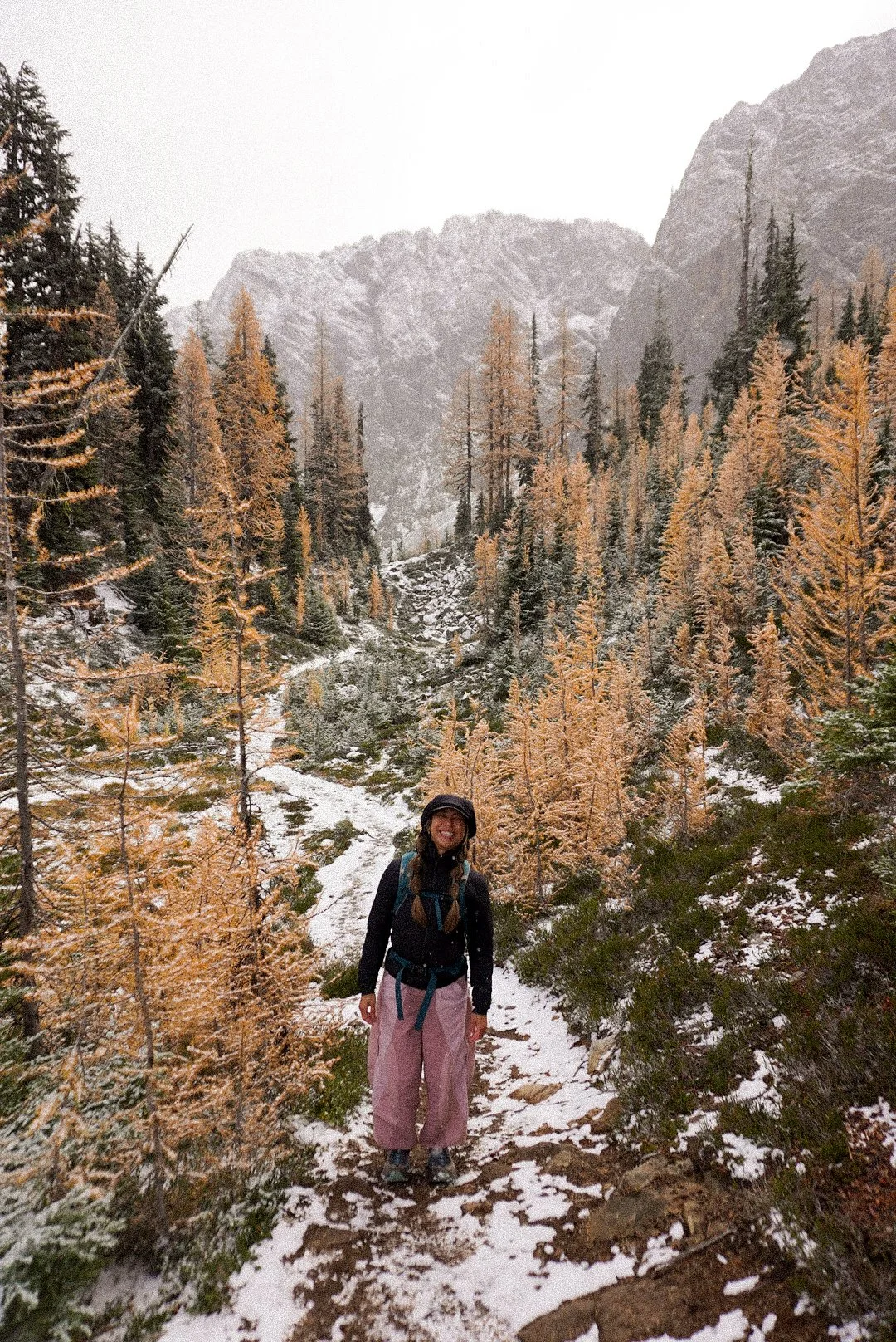 A woman hiking on a snowy trail surrounded by yellow and green trees with mountains in the background.