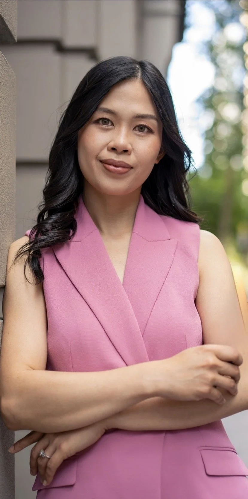 A woman with dark, wavy hair wearing a sleeveless pink blazer top, standing outdoors with trees and a building in the background.