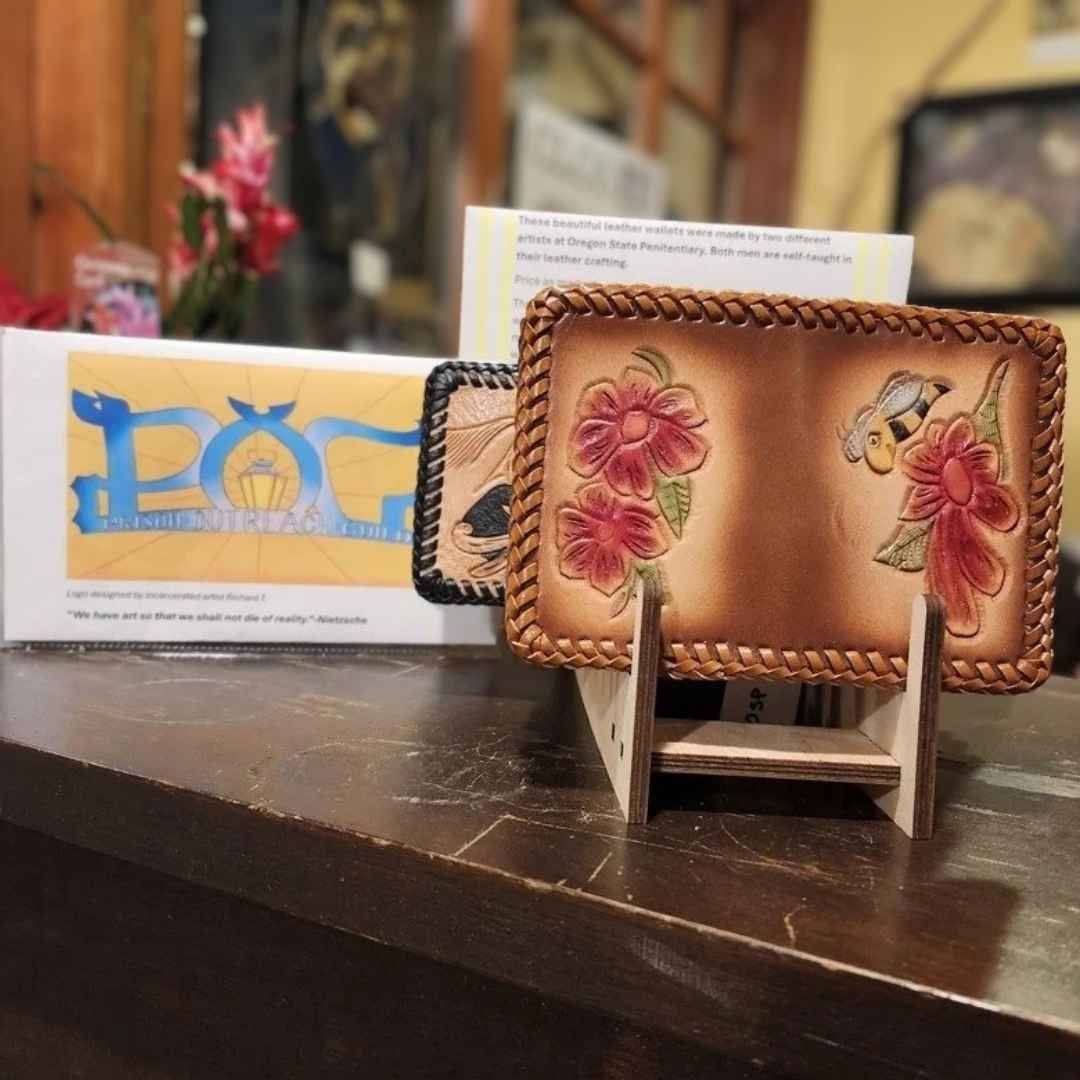 A tabletop display of prisoner-made artwork for sale, with two colorfully-tooled leather wallets in the foreground and the Prison Outreach Guild in the background.