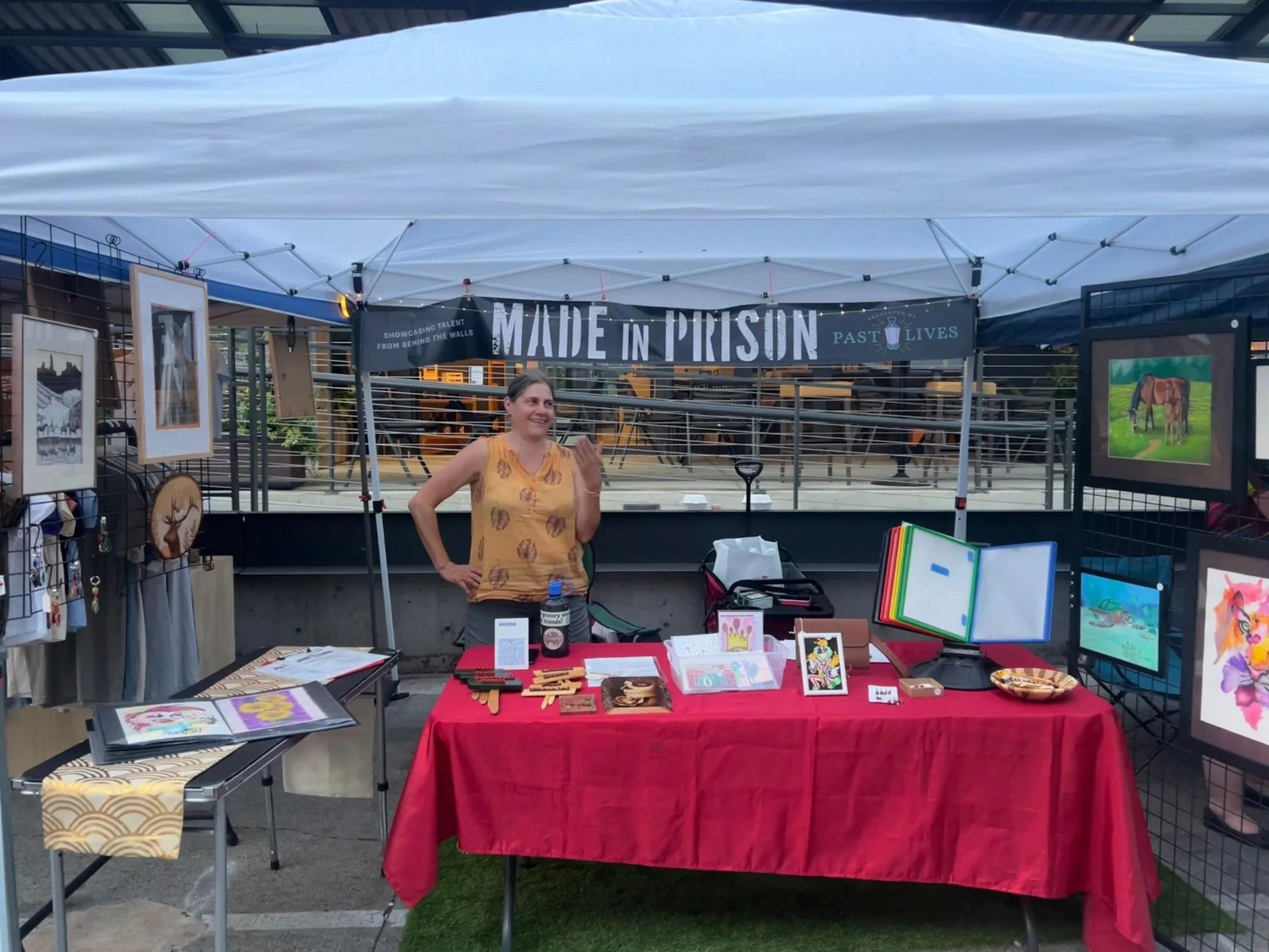 Prison Outreach Guild booth at a street market event, with a white canopy overhead and below if a table draped in red cloth, with an assortment of artworks displayed for sale.