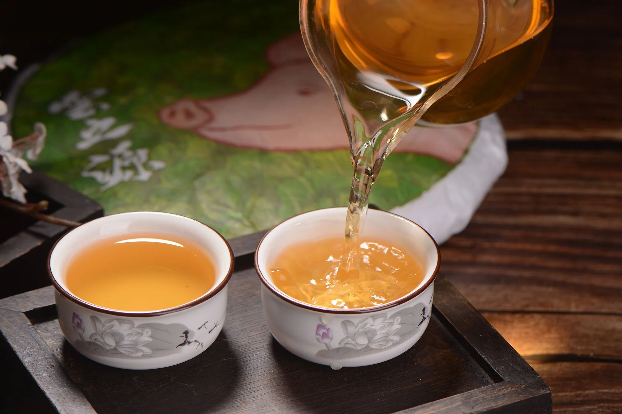 A teapot pours golden yellow tea into two cups in the foreground, and in the background is a tea cake with an illustration of a pig and gingko leaves by Bang 