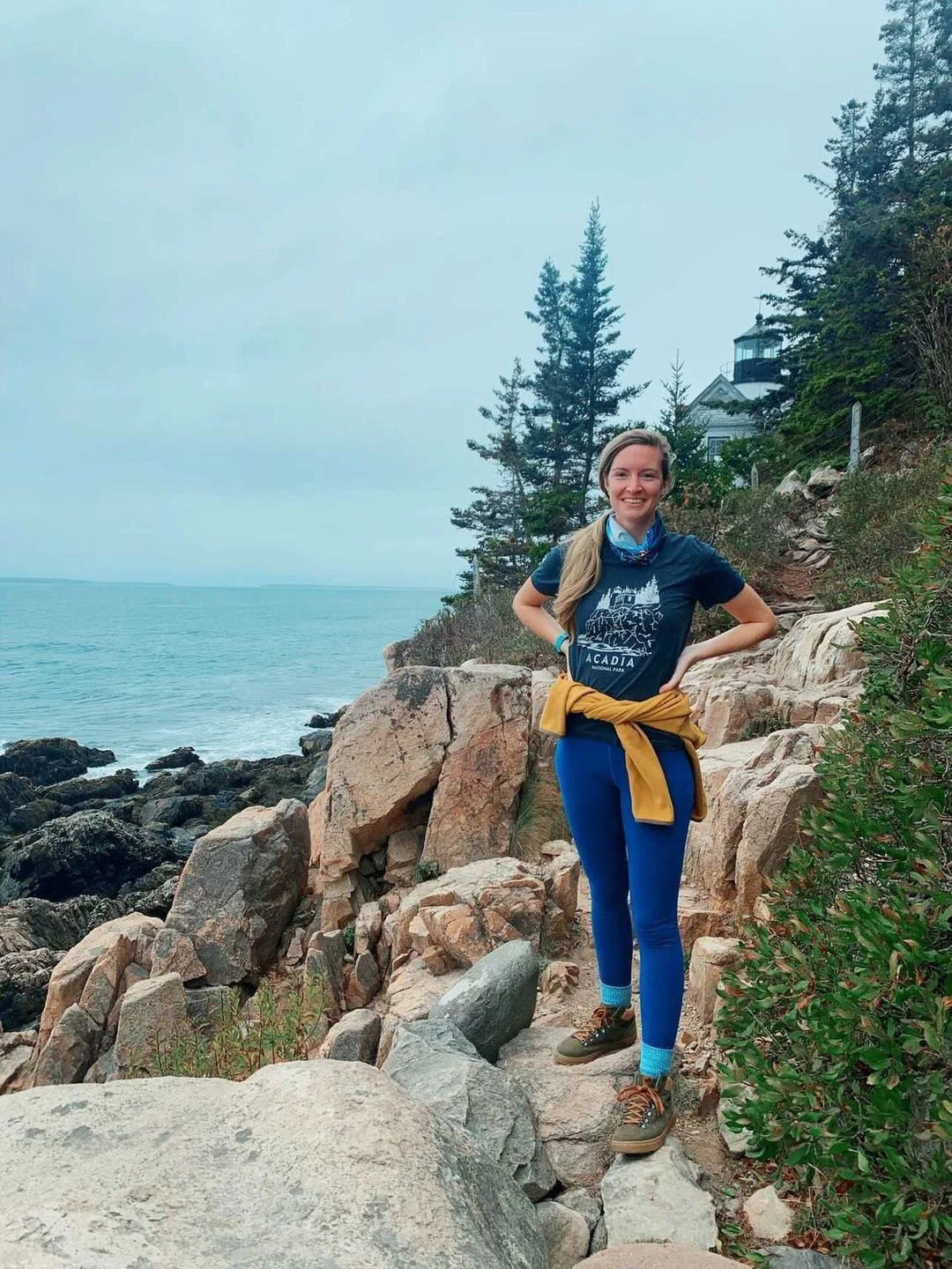 A woman stands in front of the Bar Harbor light house in Acadia national Park. She's standing next to a large pine tree on a cliff, sporting the Acadia National Park shirt designed by Bang with her hands on her hips.
