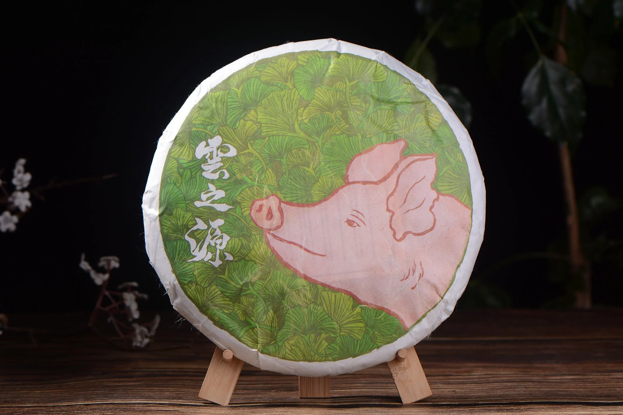 A tea cake with an illustration of a pig facing leftward, and in the background is a lush forest of ginkgo leaves. There is Chinese calligraphy on the wrapper of the tea cake