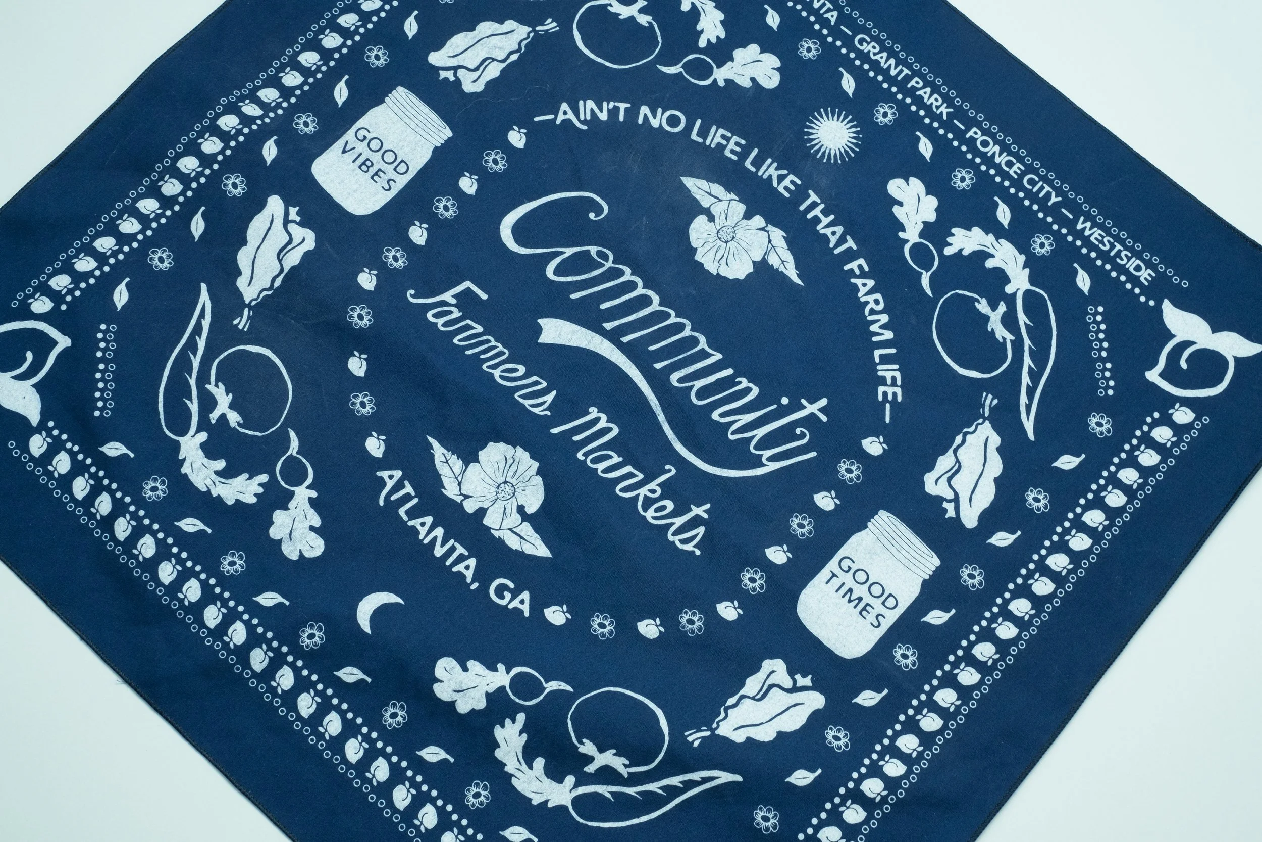 A bandana with the words Community Farmers Markets in the middle, adorned all around with elements featuring different vegetables and mason jars.