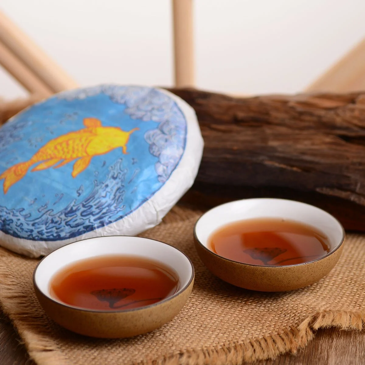 Two tea cups filled with dark brown pu erh tea in the foreground, and in the background is the tea cake illustration of the carp leaping out of the water