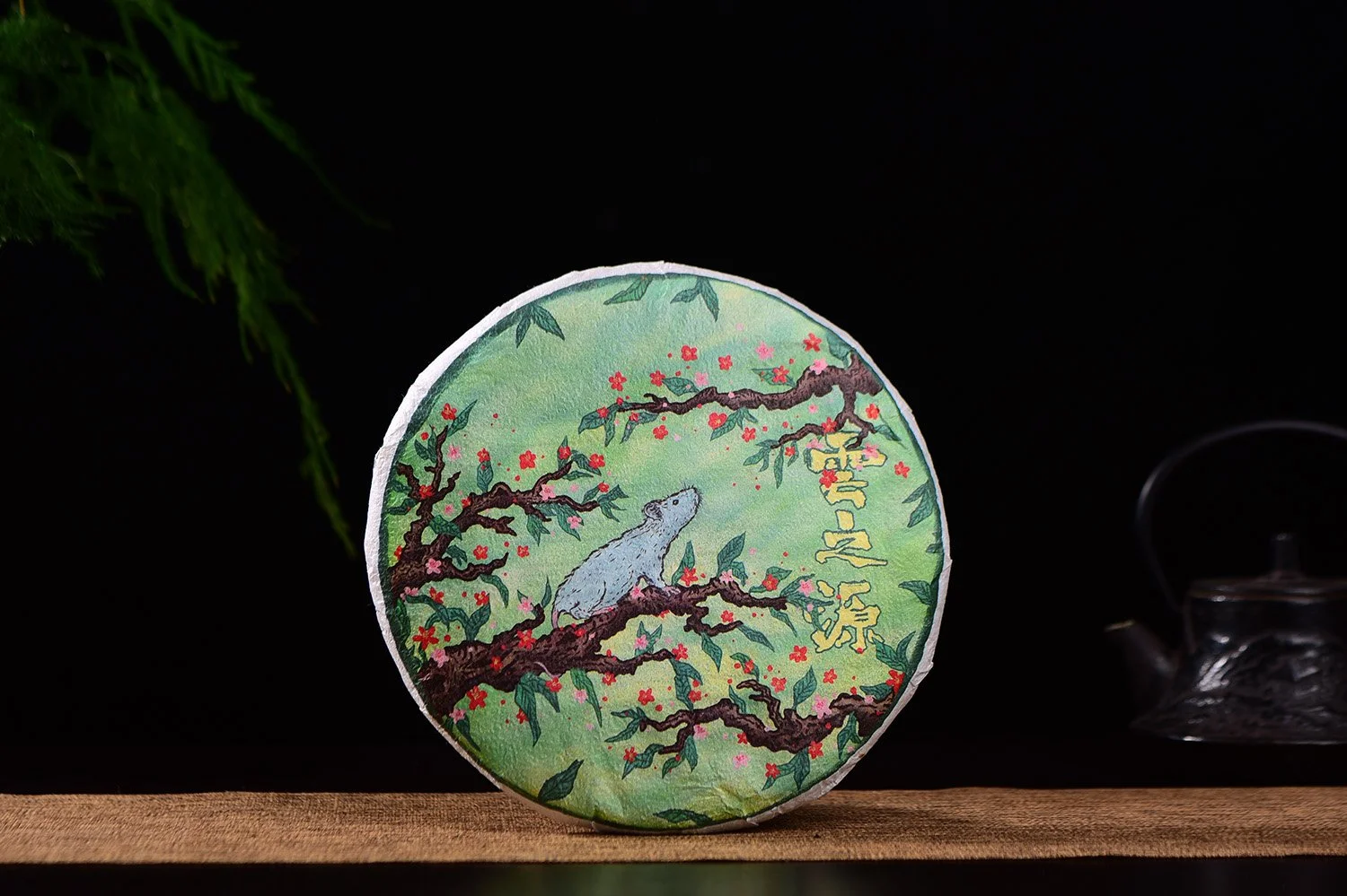 A hand illustrated rat sits on a branch filled with red and pink flowers and looks up to the right. Used as a wrapper for a Chinese tea cake 