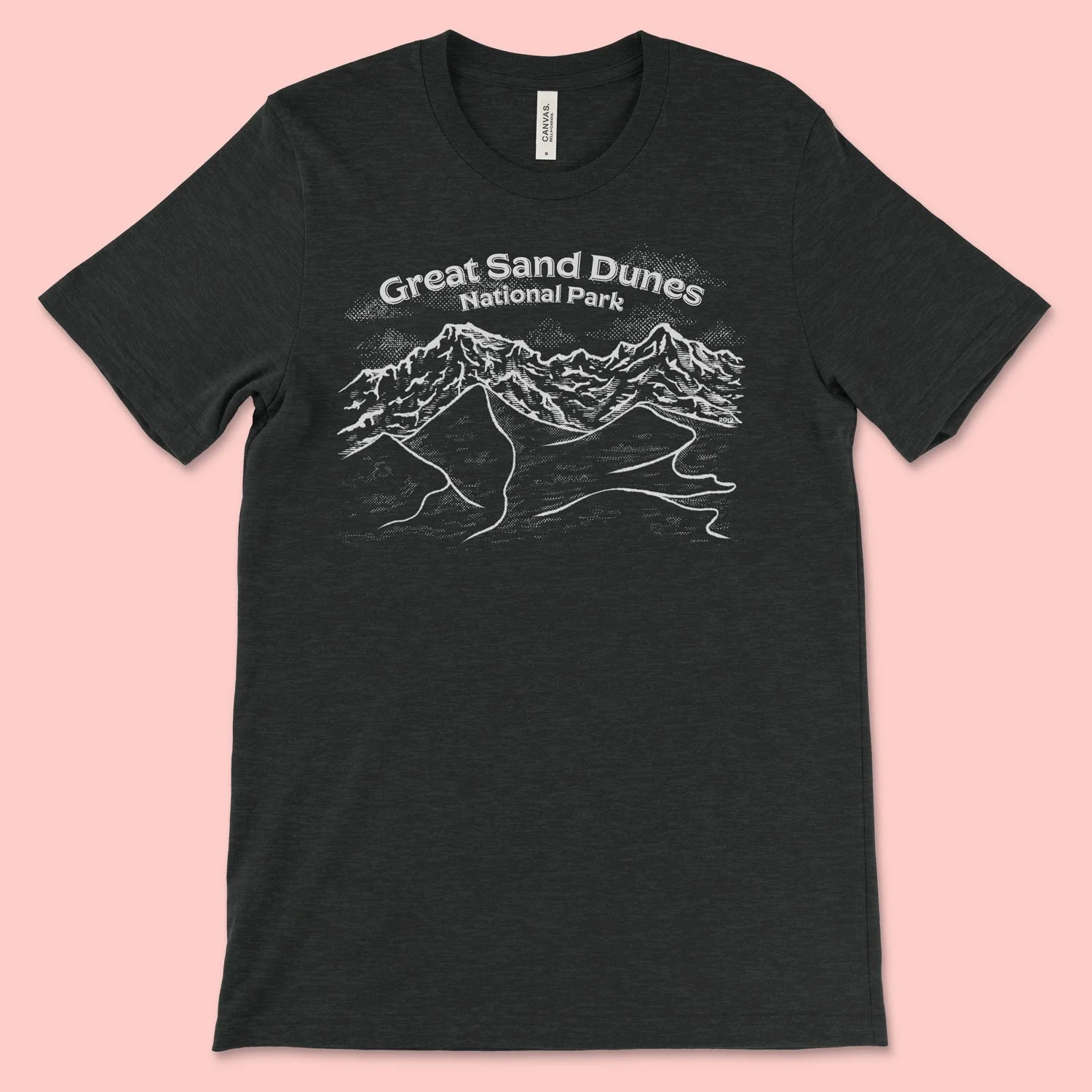 A black t-shirt featuring a one color illustration of the Great Sand Dunes National Park in Colorado. Large dunes of sand sit in front of a backdrop of mountains, and it's designed in a way reminiscent of vintage postcards
