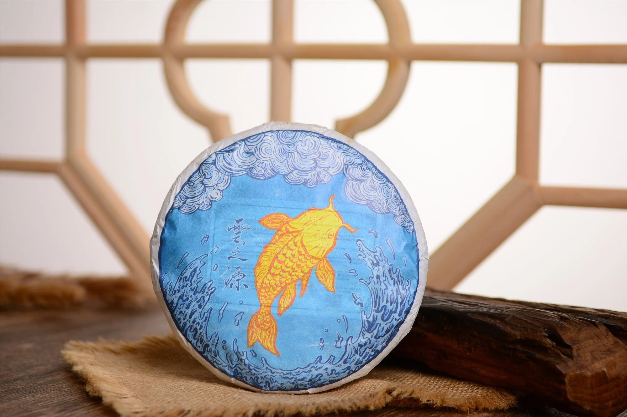 A tea cake illustration depicting a golden carp splashing upward amid a river, reaching towards swirling grey and blue clouds. It references an old Chinese folktale of carp turning into dragons once they swim up waterfalls