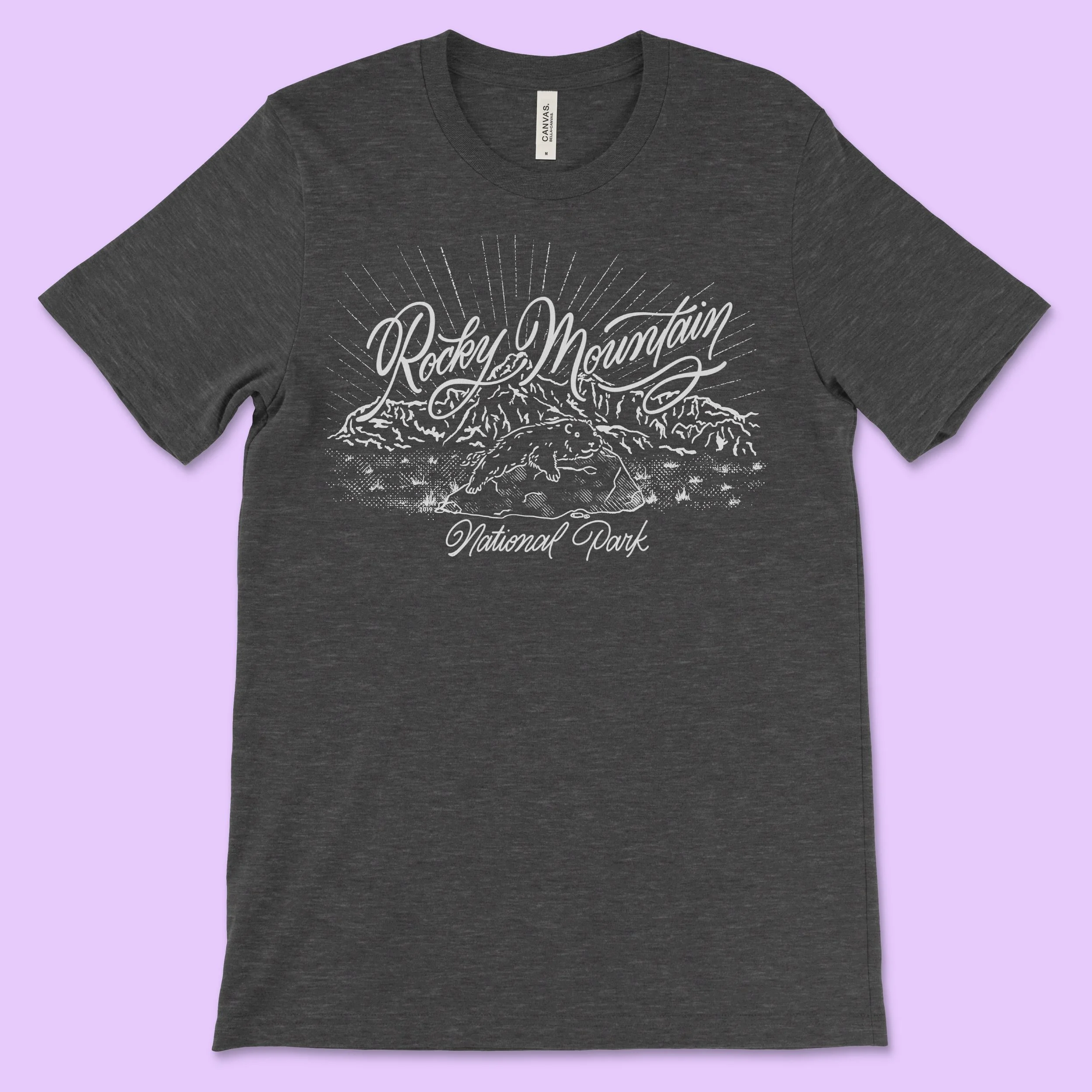 A gray shirt depicting a marmot sitting on a rock in in a field in front of a mountain range, and Rocky Mountain National Park is written across the shirt in calligraphy. The shirt is designed in one color and is reminiscent of vintage postcards 