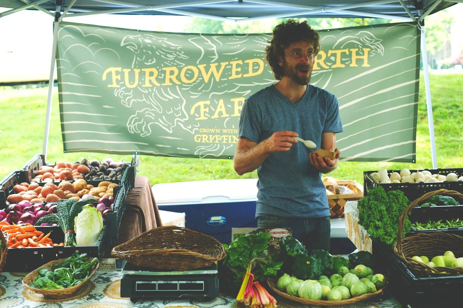 A farmer stands in front of a large banner featuring the text Furrowed Earth Farm, and a large griffin is also on the banner. The farmer is surrounded by vegetables.