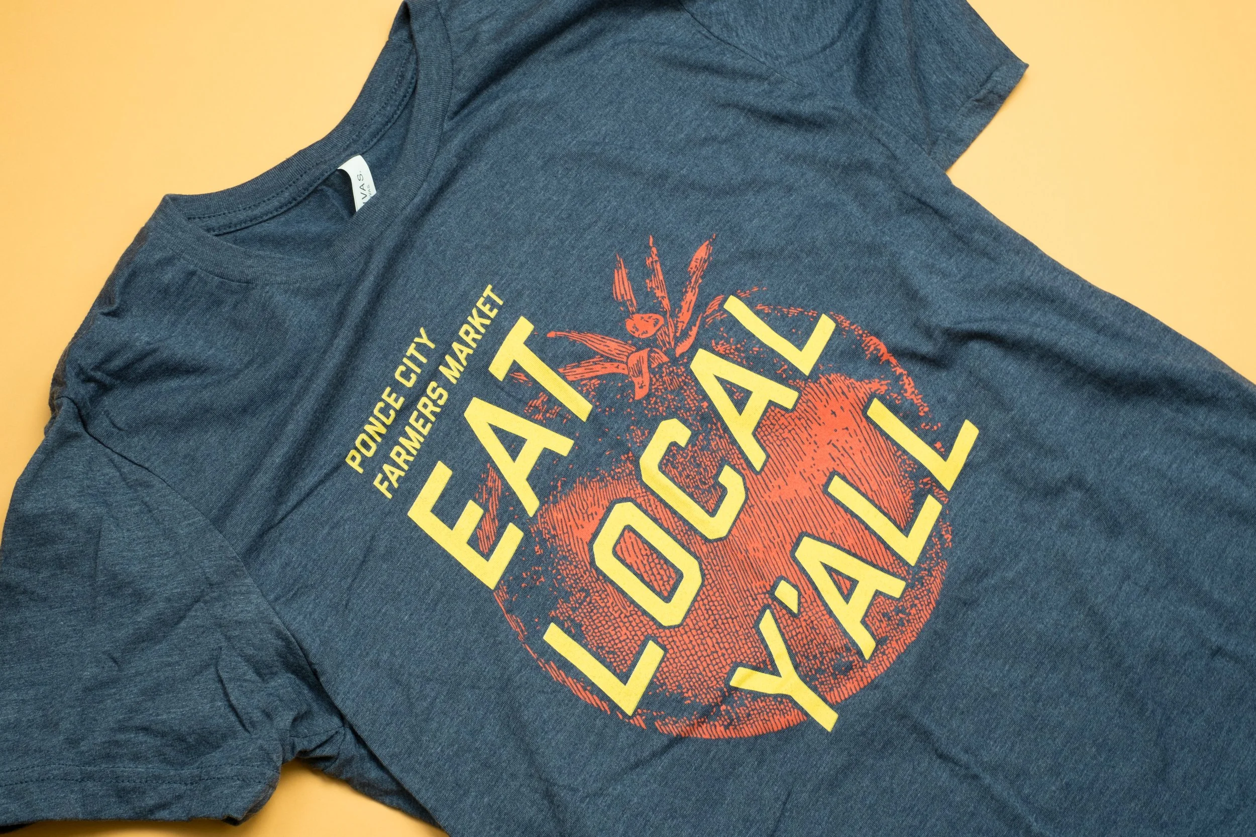 A close up of a navy blue shirt with the print Ponce City Farmers Market and yellow text saying Eat Local Y'all in front of a vintage illustration of a tomato
