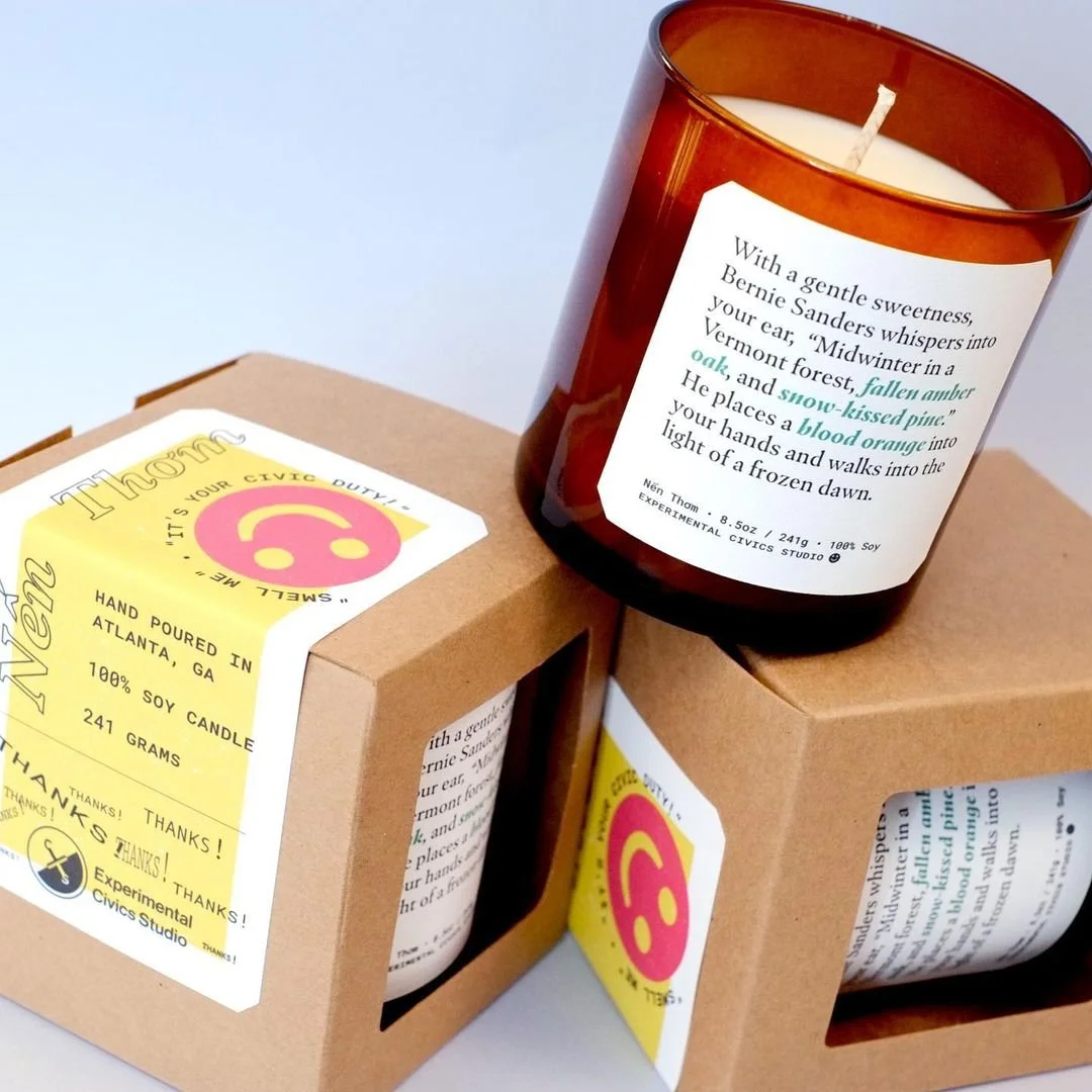 3 candles sit on top of each other. The label on the candle reads, "With a gentle sweetness, Bernie Sanders whispers into your ear, 'Midwinter in a Vermont forest, fallen amber oak, and snow-kissed pine. He places a blood orange into your hands and w