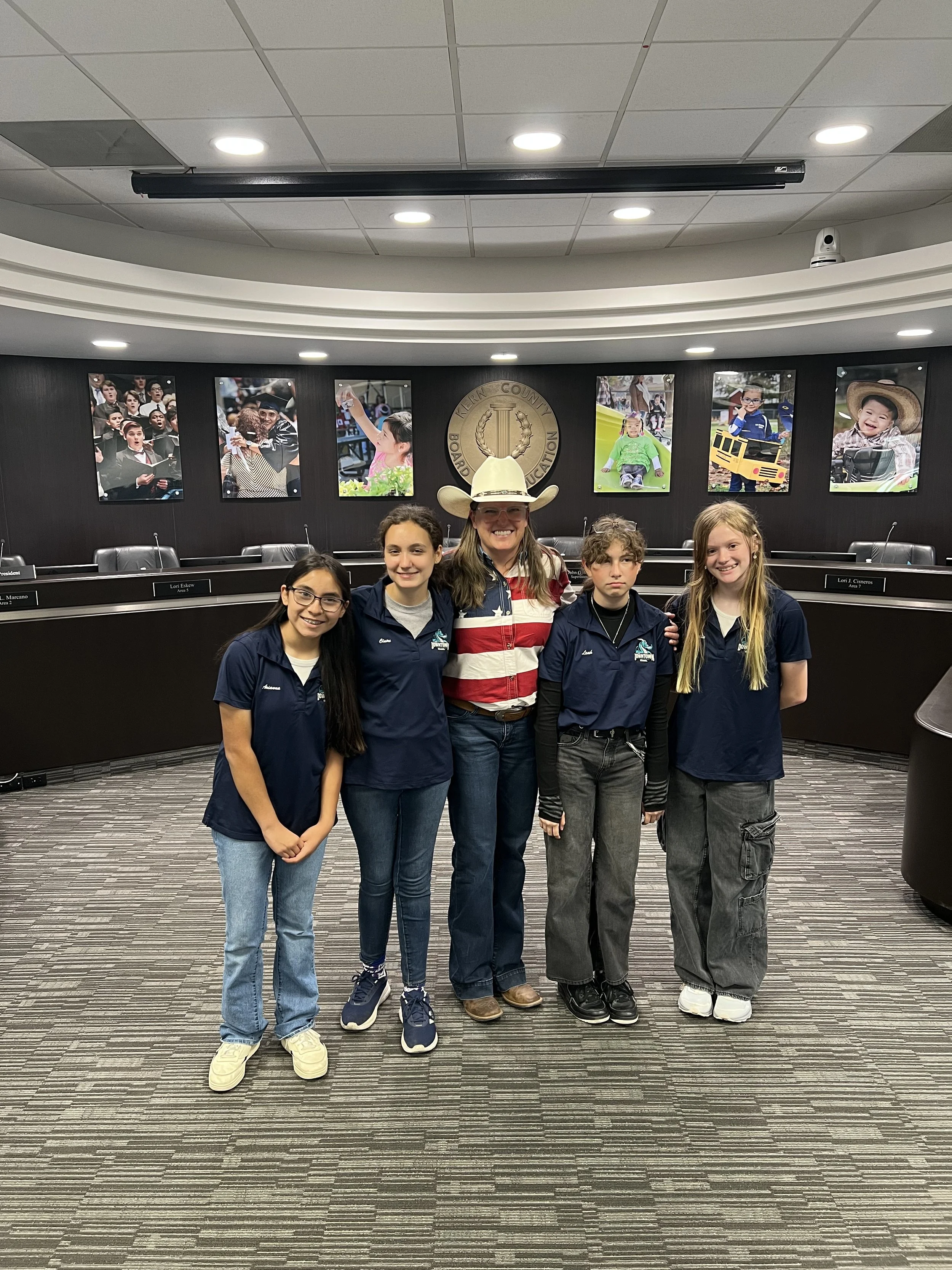 A group of five children and one adult woman standing together in a meeting room with a black wall, photographs of people, and the County Board of Education logo in the background.