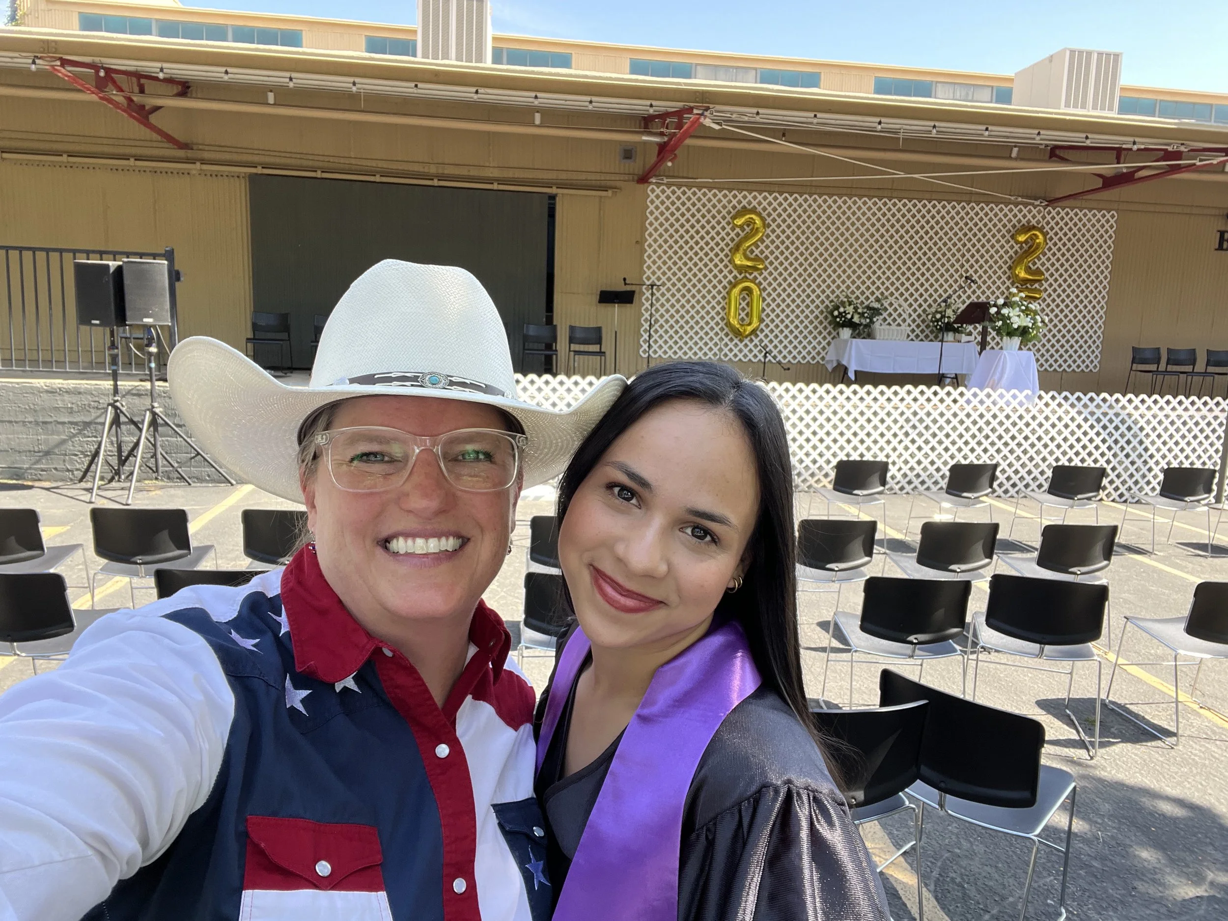 Two women taking a selfie at an outdoor event, one wearing a cowboy hat and glasses, the other with long dark hair. In the background, there are chairs arranged for an audience, a stage with balloons that spell out "2023," and tables with flowers and decorations, set against a building with a yellow exterior and large windows.