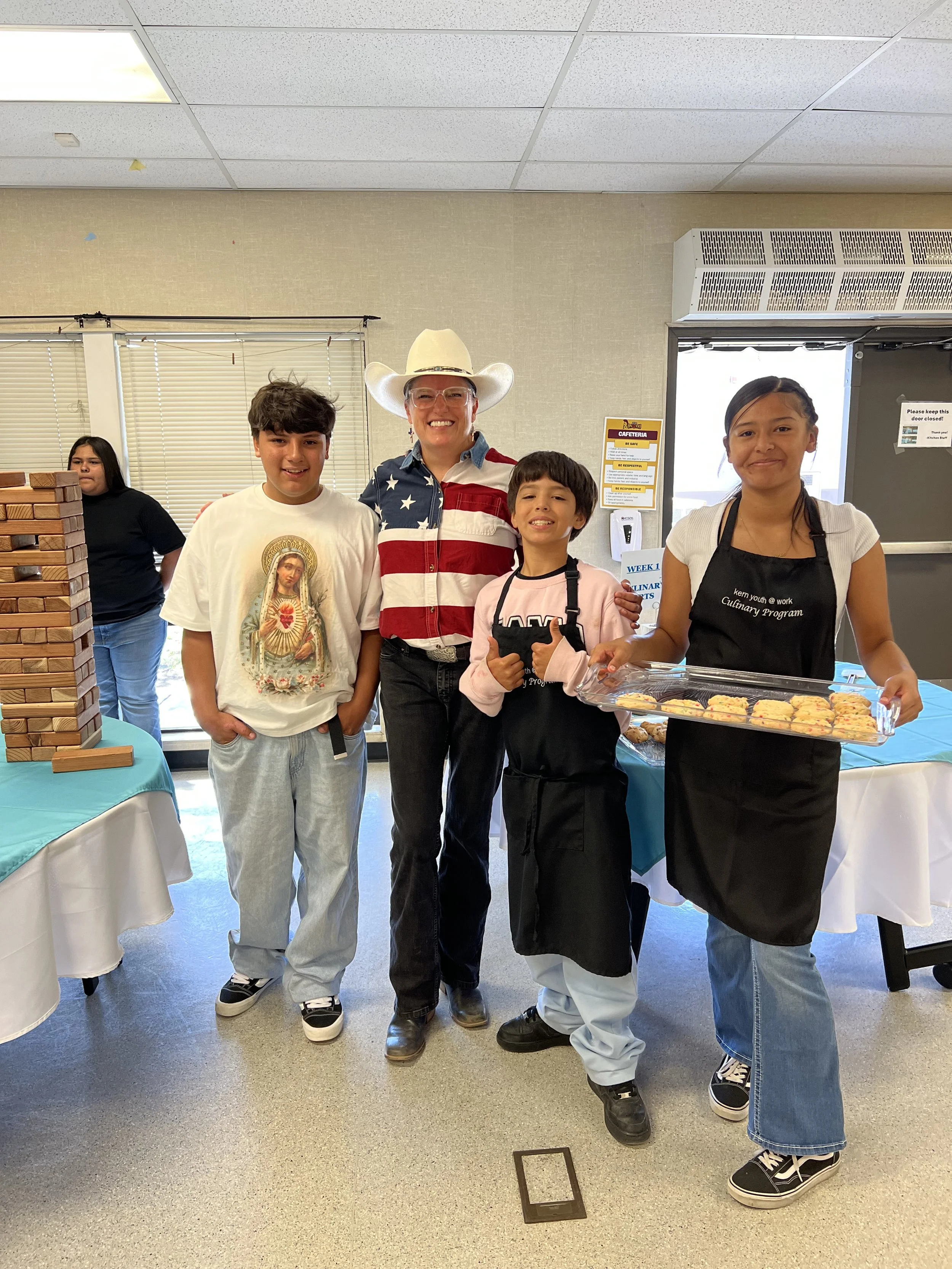 Group of five people standing indoors, with a man wearing a white cowboy hat and a patriotic shirt in the center, two boys and two girls surrounding him, one girl wearing a black apron and holding a tray of baked goods, and a woman in the background behind a table, with a Jenga game on the left.