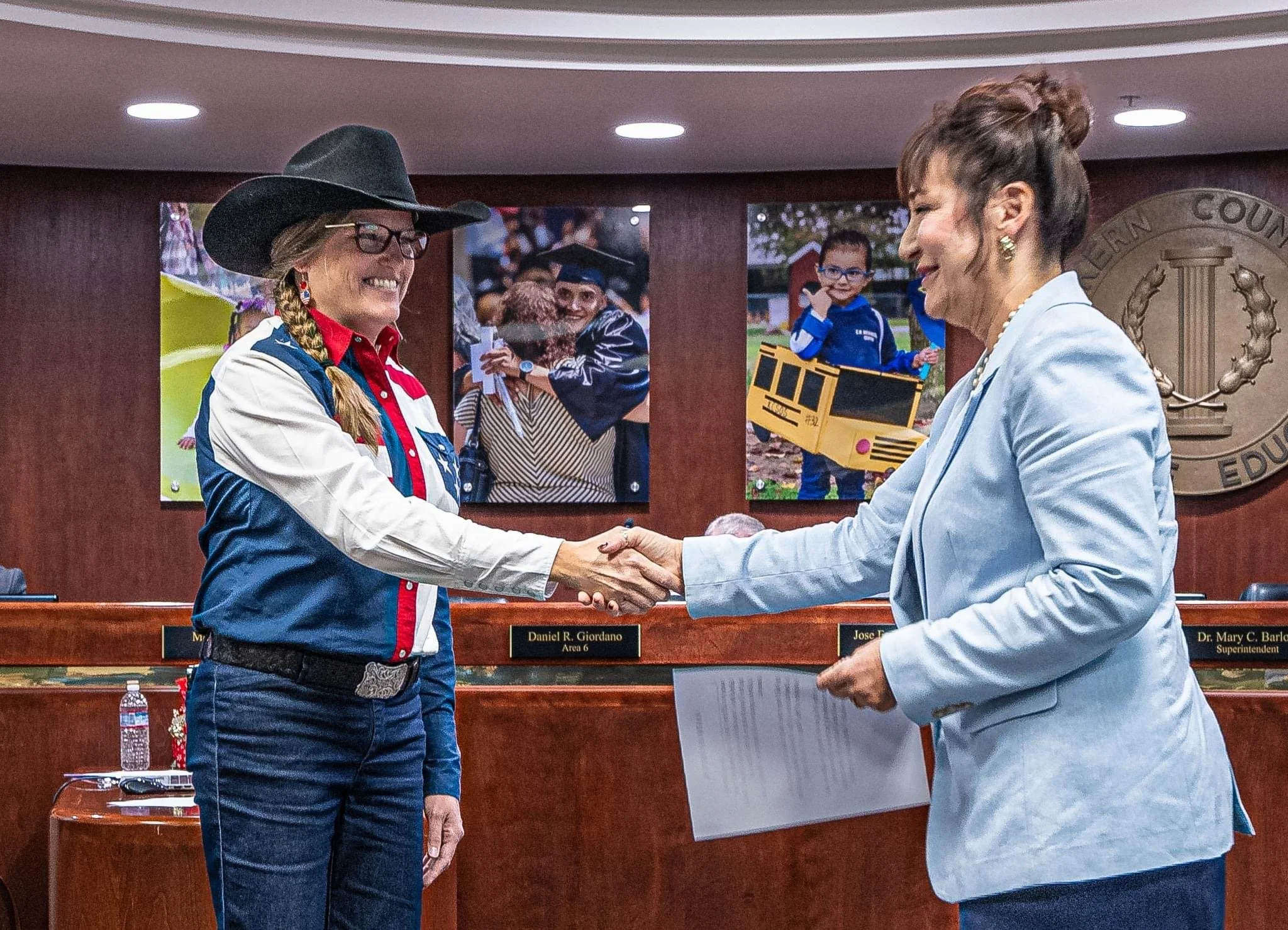 Two women shake hands in an award or recognition ceremony, smiling at each other. Behind them, there are photographs of children and a statue or emblem of a seal on the wall.