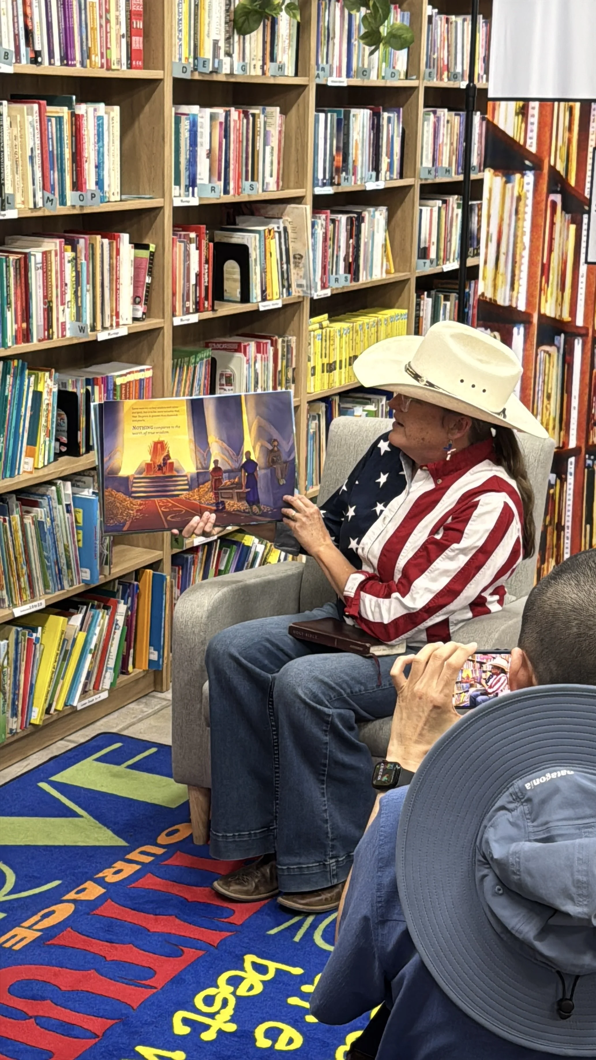 A woman wearing a cowboy hat and patriotic shirt reading a picture book to children in a library. The background shows shelves filled with children's books, a blue educational rug on the floor, and people taking photos of her.