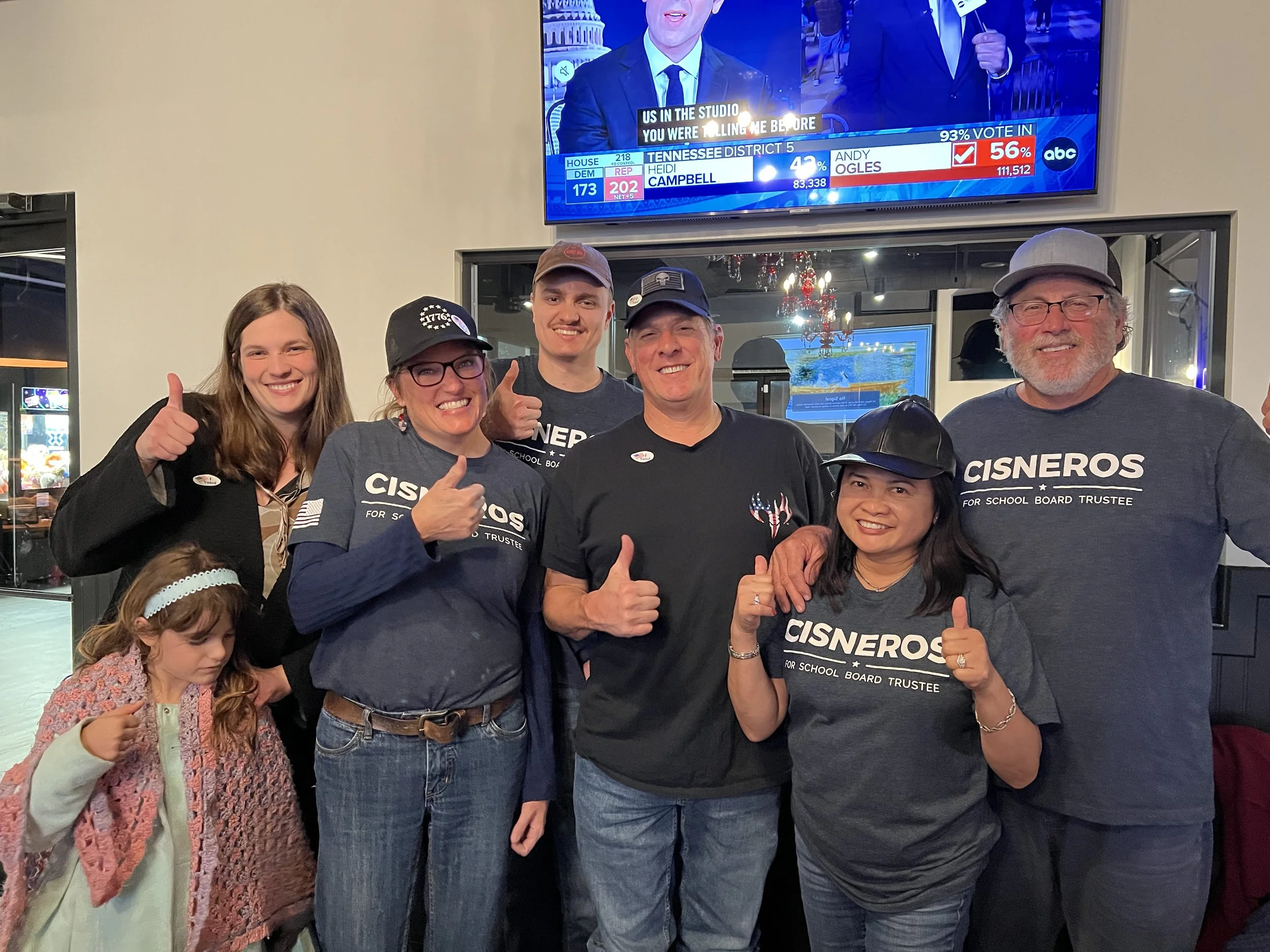 A group of six diverse people smiling and posing for a photo. Four adults and two children, some giving a thumbs-up. The background shows a television screen displaying election results and a reflection of a chandelier.