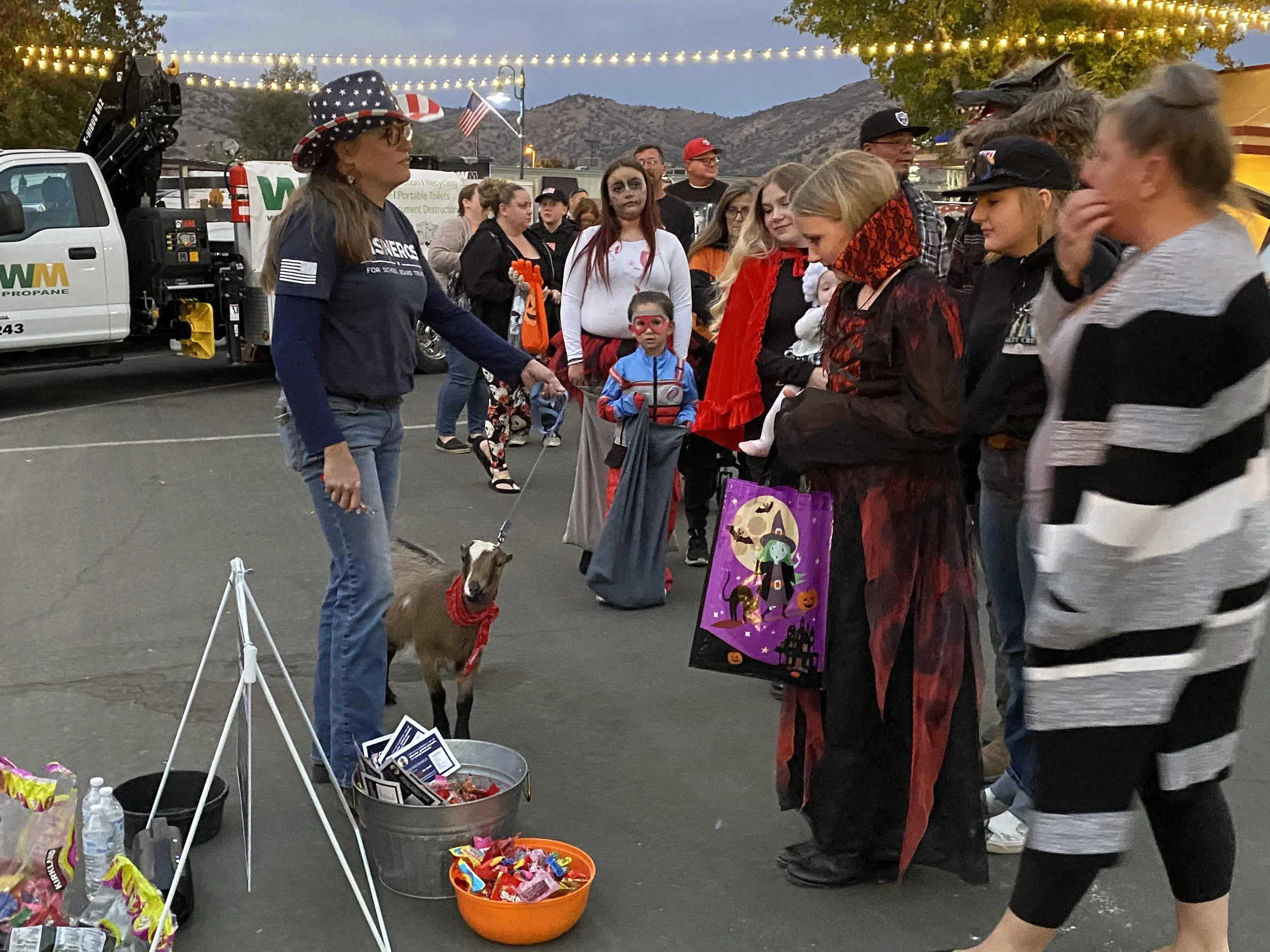 People dressed in Halloween costumes gathering around a street vendor's booth at dusk. The group includes a woman with a dog on a leash, children, and adults in various costumes. A truck with propane equipment and string lights are visible in the background.