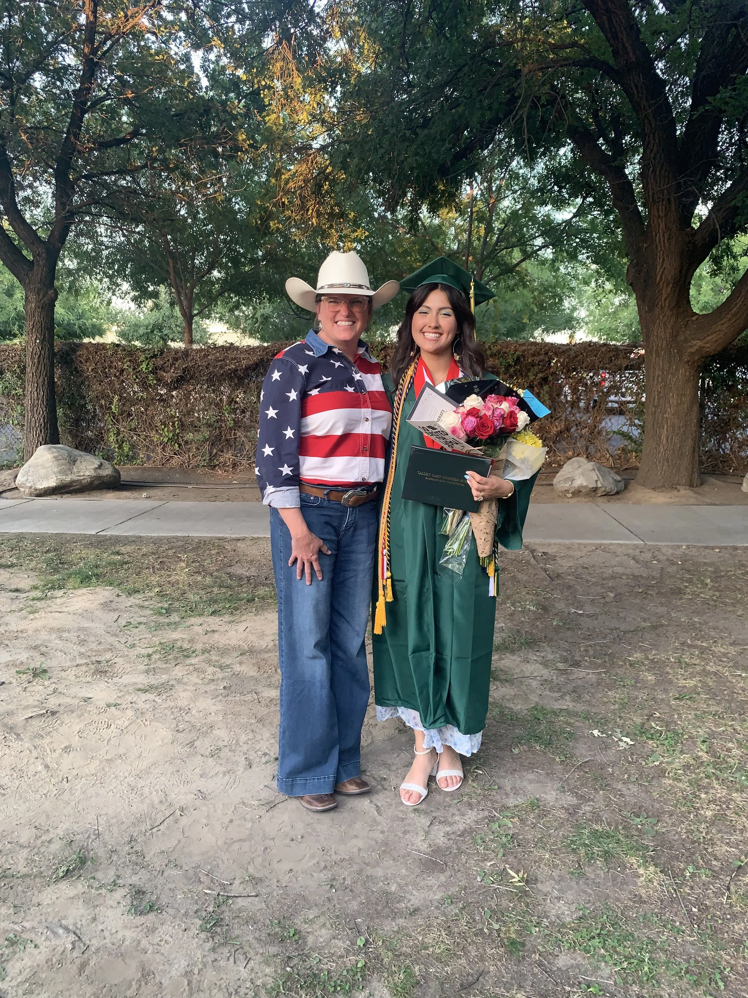 A woman in a green graduation gown and cap holding a bouquet of flowers, standing next to a woman wearing a cowboy hat and an American flag-themed shirt, both smiling outdoors with trees and rocks in the background.