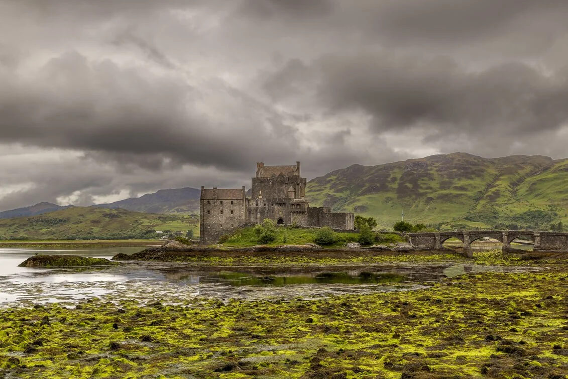 Eilean Donan Castle in the Scottish Highlands surrounded by tidal shoreline and green hills under dramatic cloudy skies
